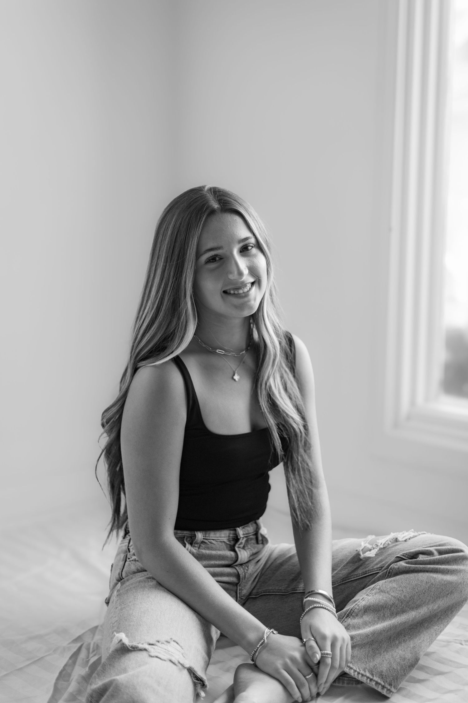 Young woman in black top and light jeans smiling while sitting casually indoors.