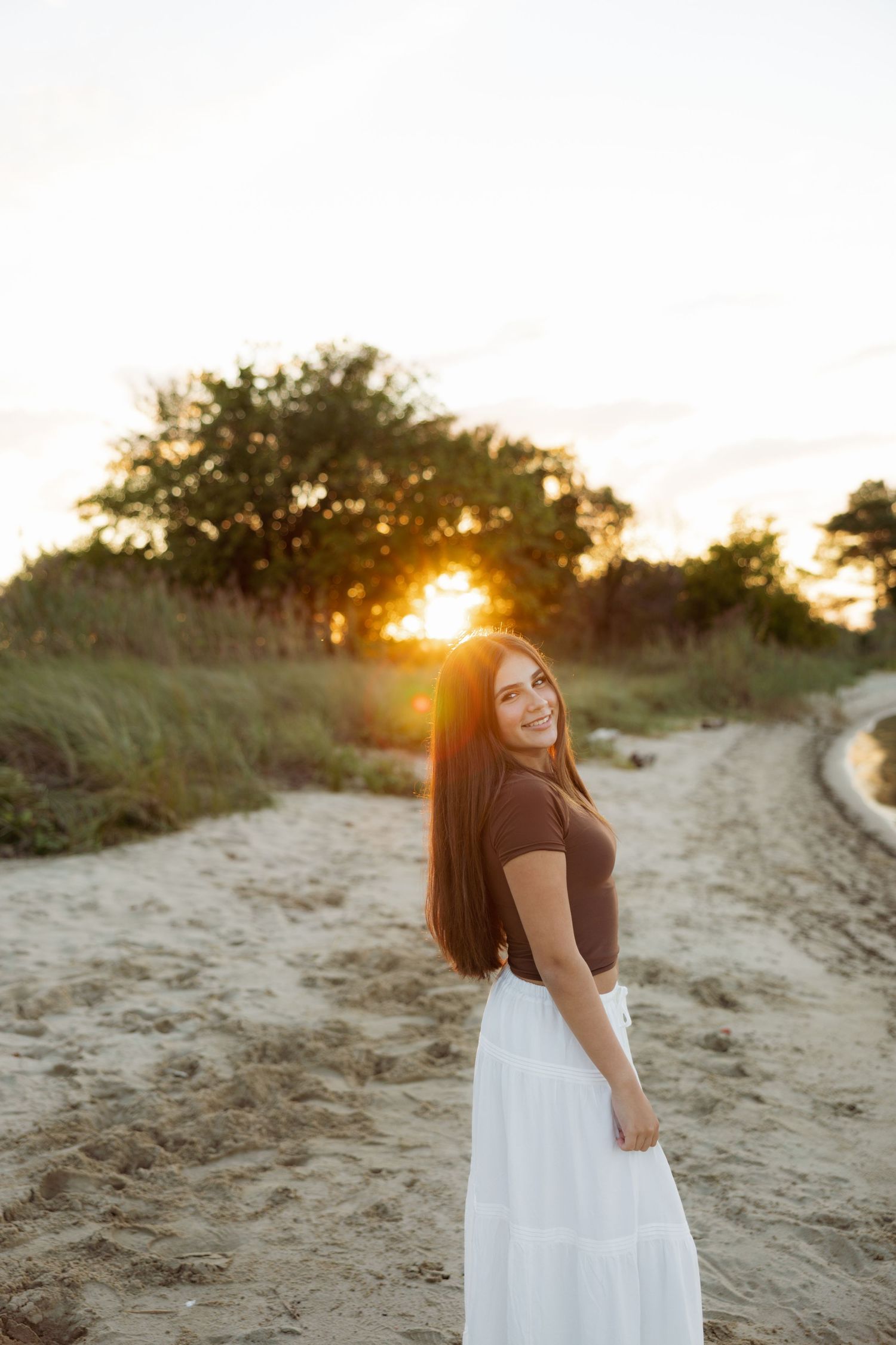 A person in a white skirt and dark top poses against a sandy beach path at golden hour sunset.