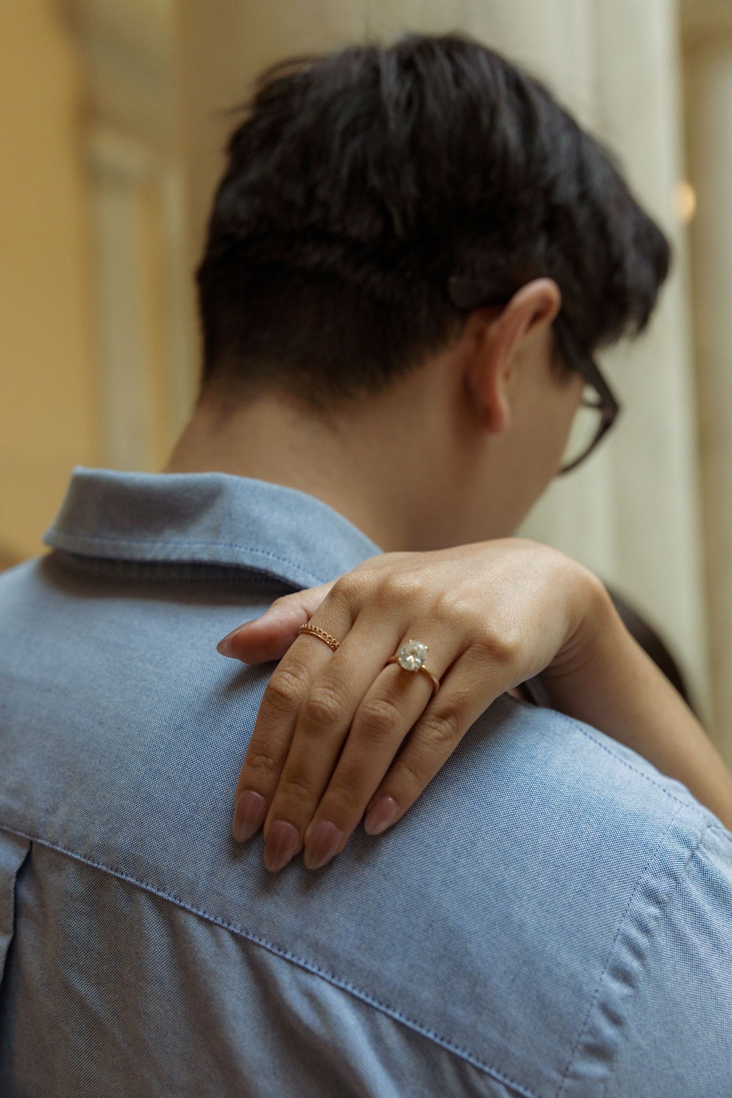 A woman's hand with a diamond engagement ring rests on a man's shoulder in a blue denim shirt.