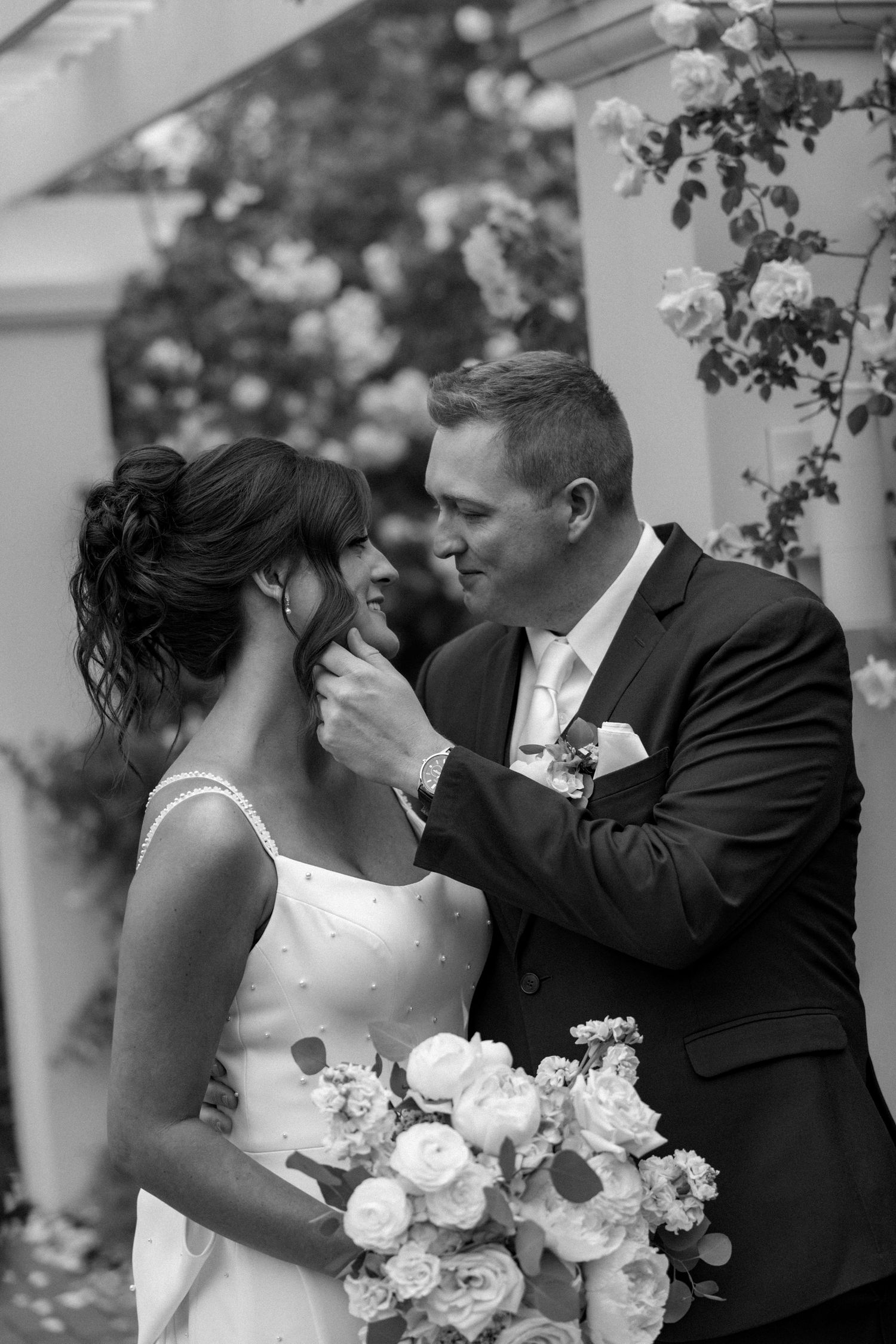Black and white wedding photo of groom tenderly touching brides face, surrounded by roses.