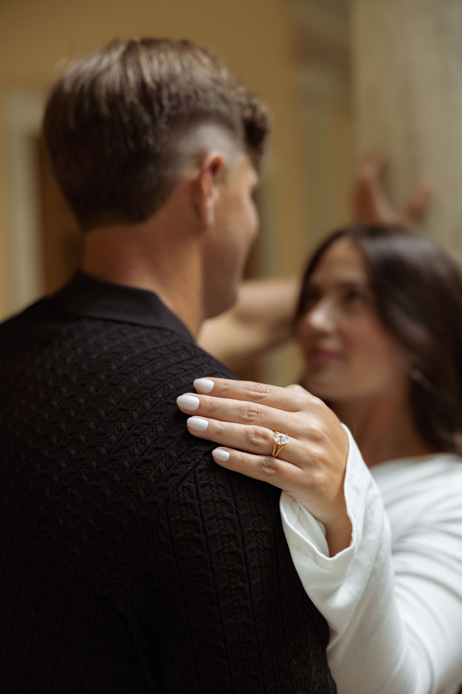 Couple embracing closely, woman wearing a gold engagement ring with white manicure resting her hand on man's black sweater.