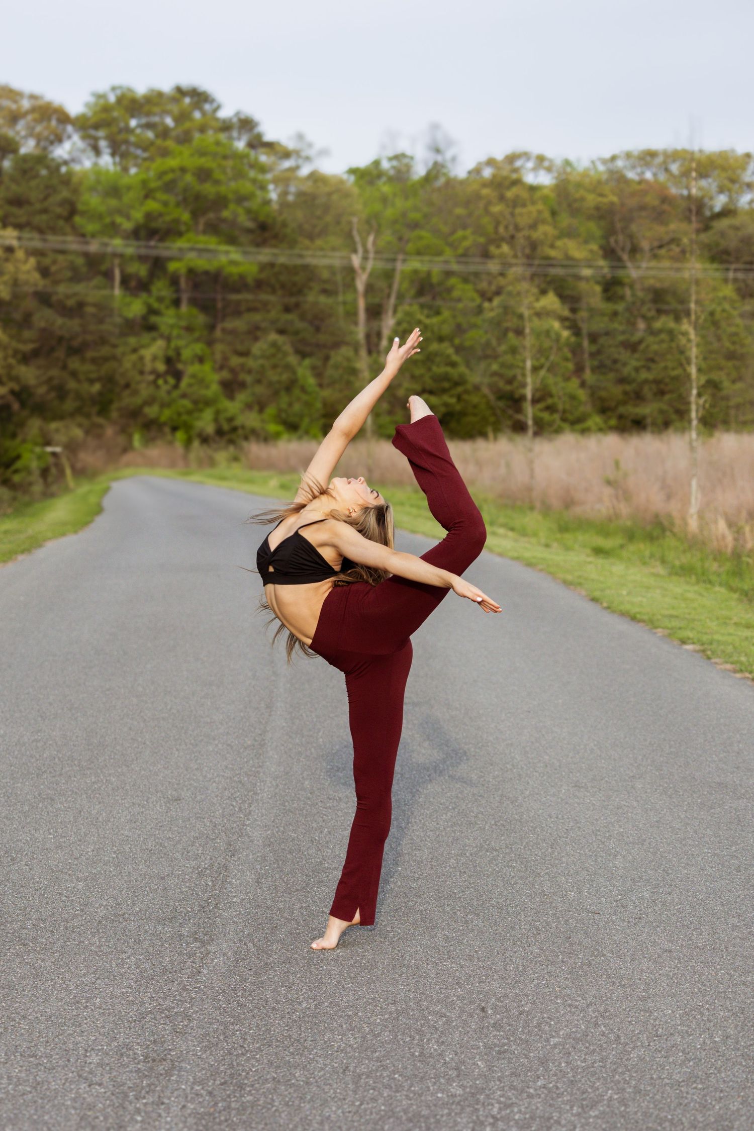 Athletic dancer executes high kick pose on rural road wearing burgundy outfit.