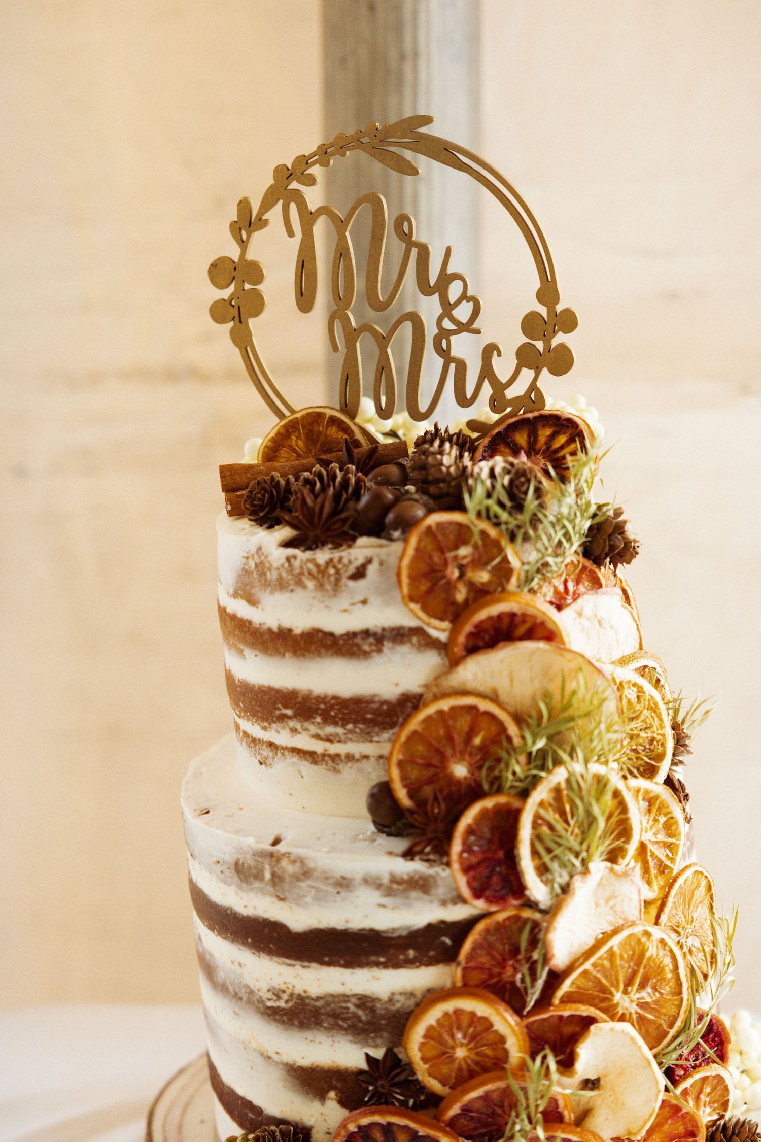 Two-tier naked wedding cake with dried citrus, pine cones, and a gold Mr & Mrs topper.