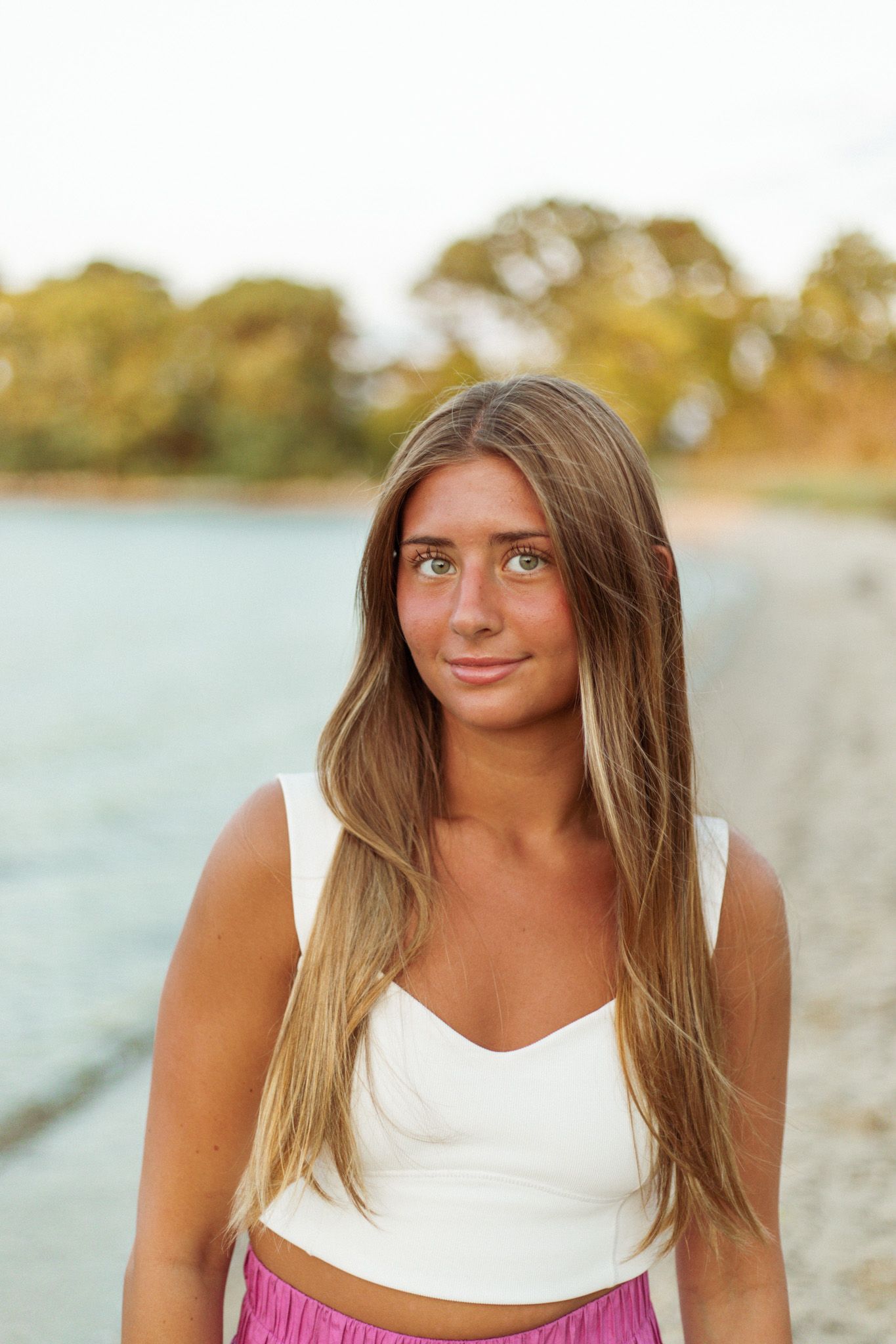 Person in white top and pink skirt poses near water at sunset.