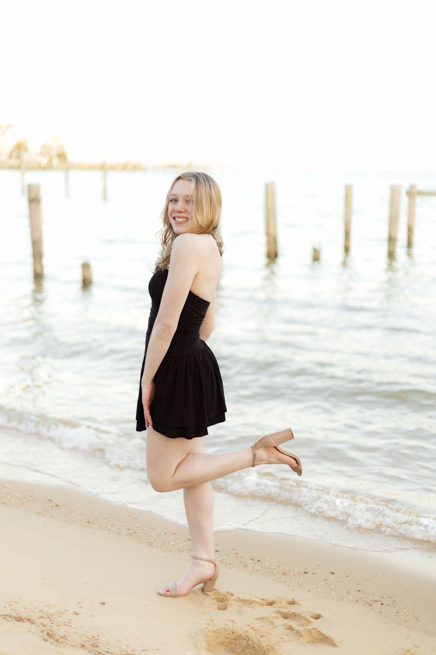 Person in black dress poses playfully on beach near wooden posts.