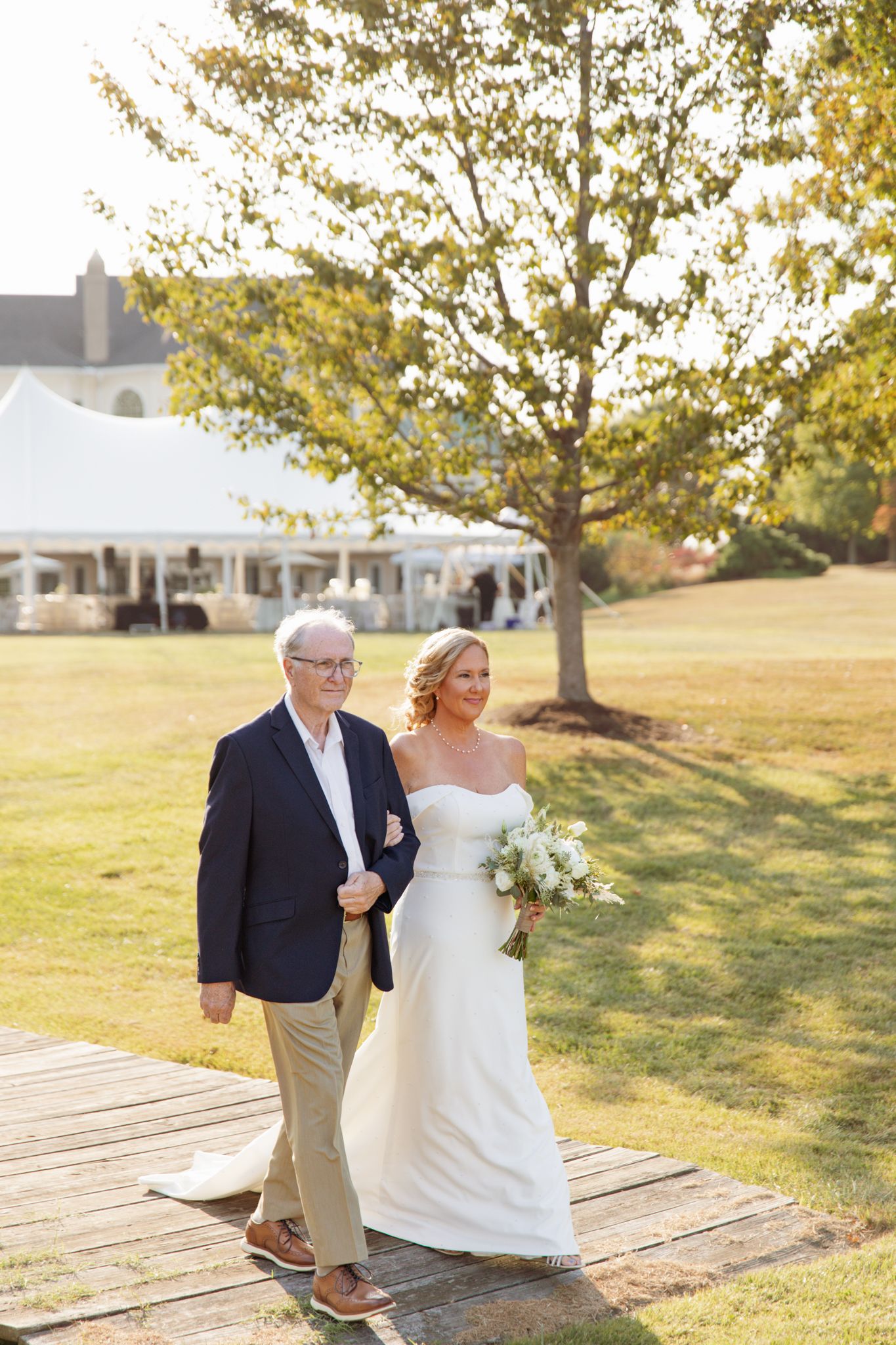 Bride in white strapless gown holding bouquet walks arm-in-arm with older man in navy blazer on wooden path outdoors.