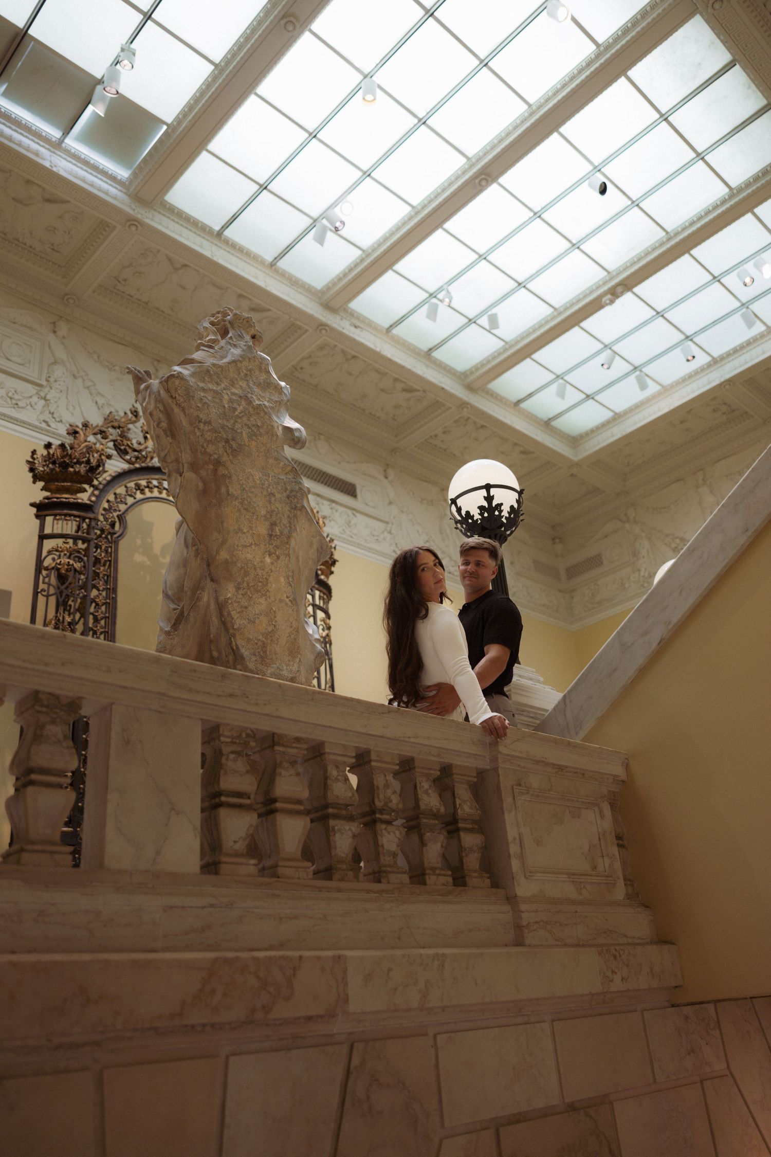 Ornate marble staircase with decorative balustrades and crown moldings illuminated by natural light through a glass ceiling.