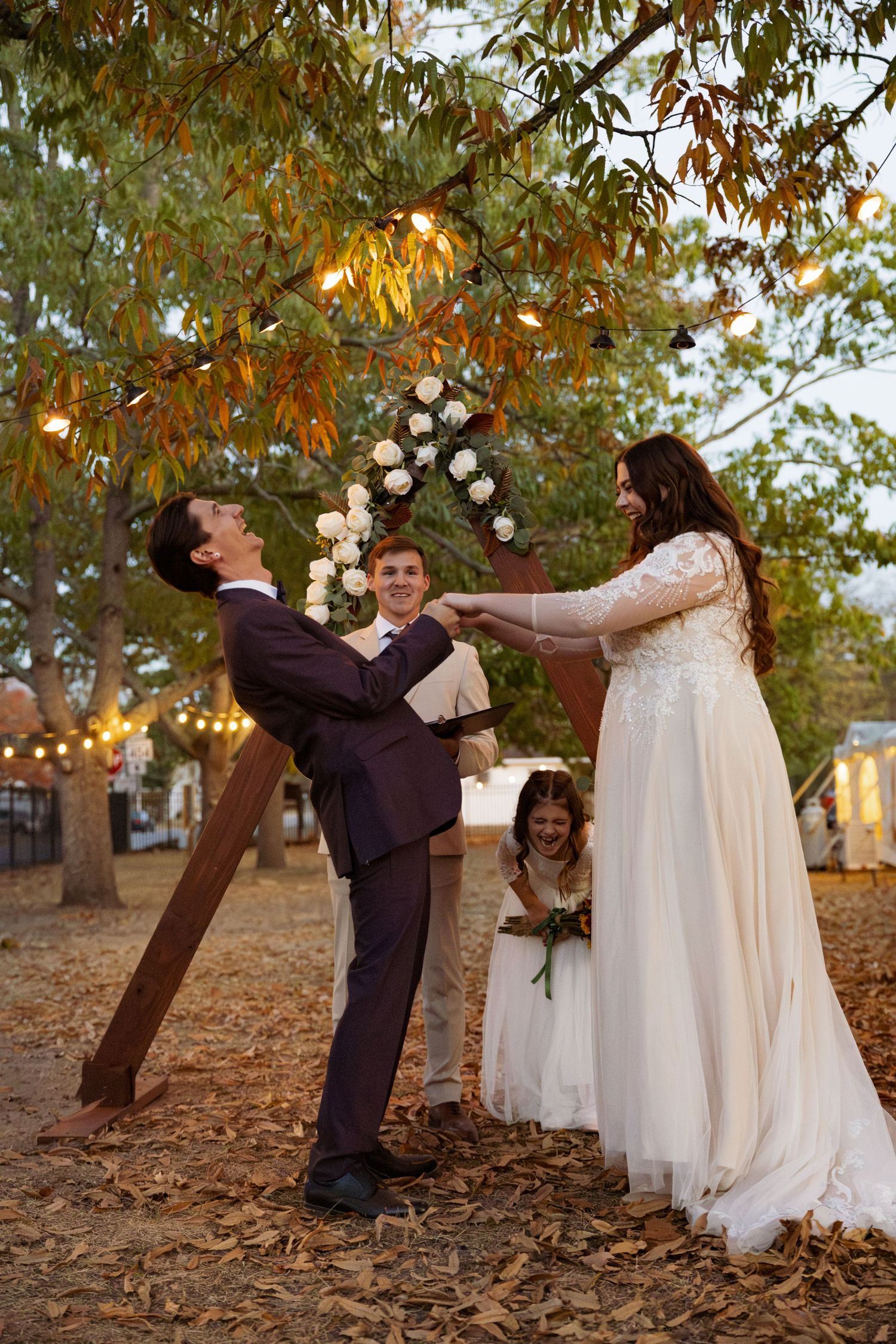 Bride and groom share a joyful moment outdoors during wedding ceremony with floral arch, string lights, and fall leaves.