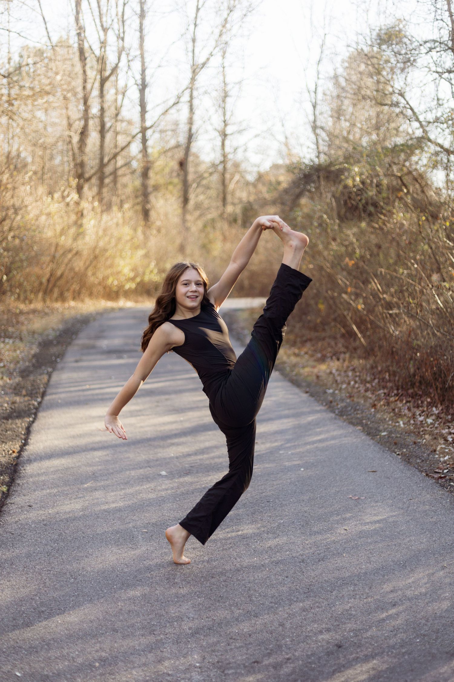 Dancer performs high kick on autumn path surrounded by golden trees.