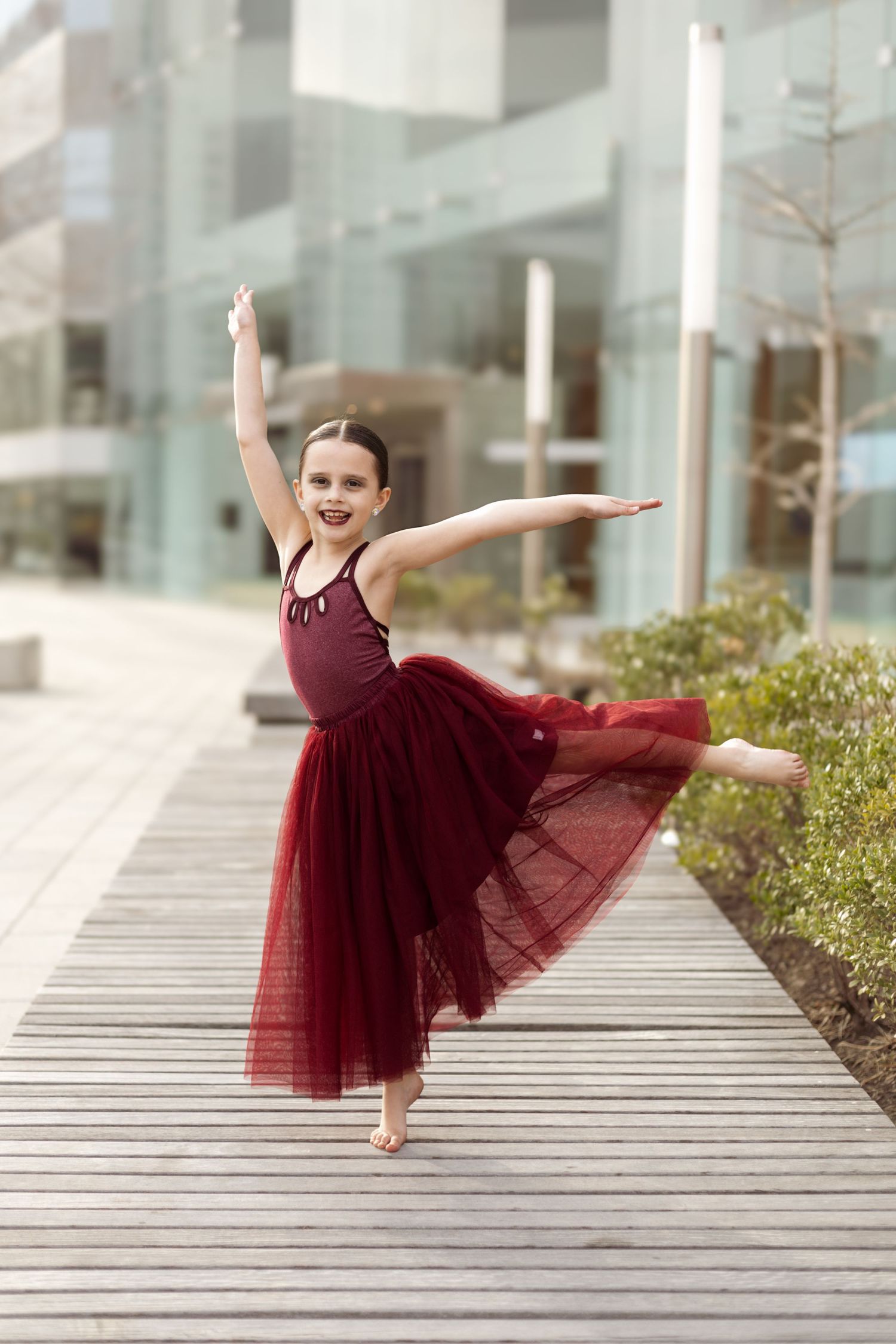 Dancer in burgundy tulle dress performs elegant pose on wooden boardwalk.