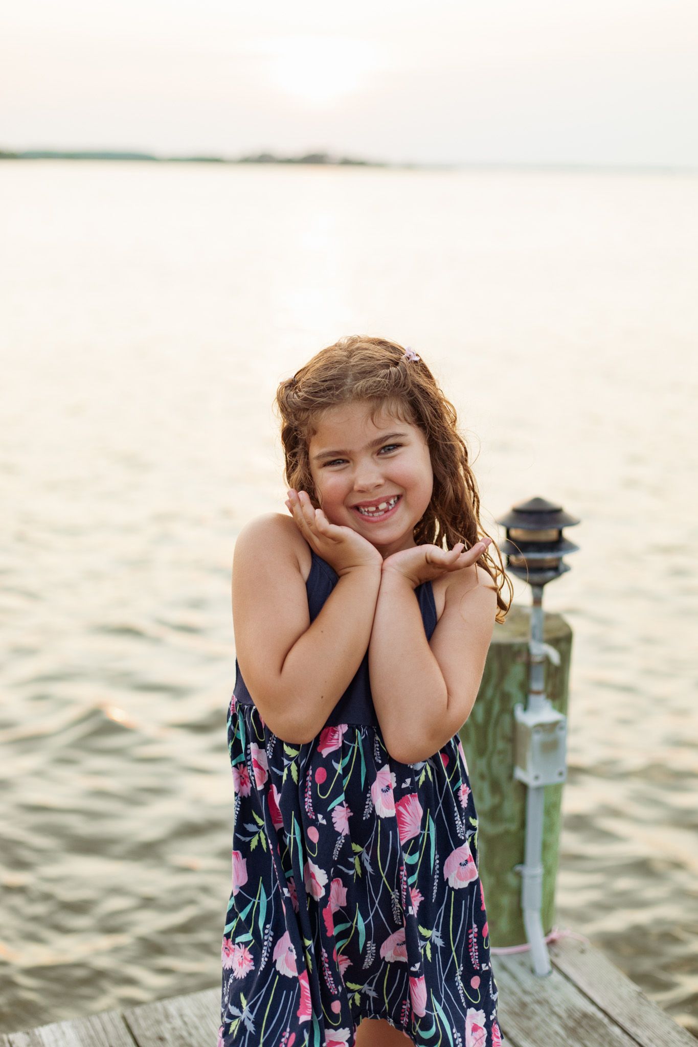 Young girl in floral dress poses cheerfully on wooden dock at sunset.