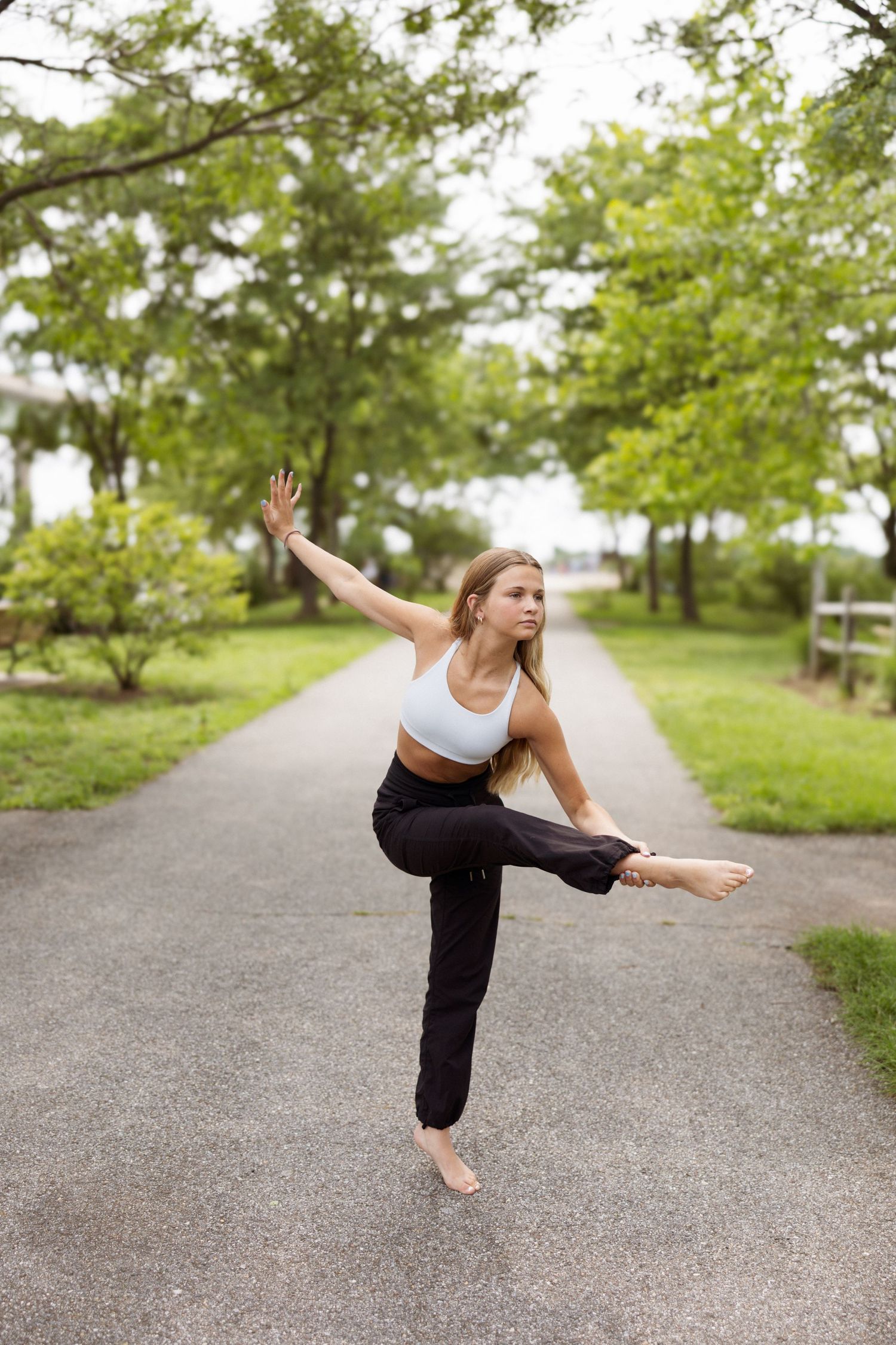 A dancer performs a graceful yoga sequence on a tree-lined path during an outdoor practice session.