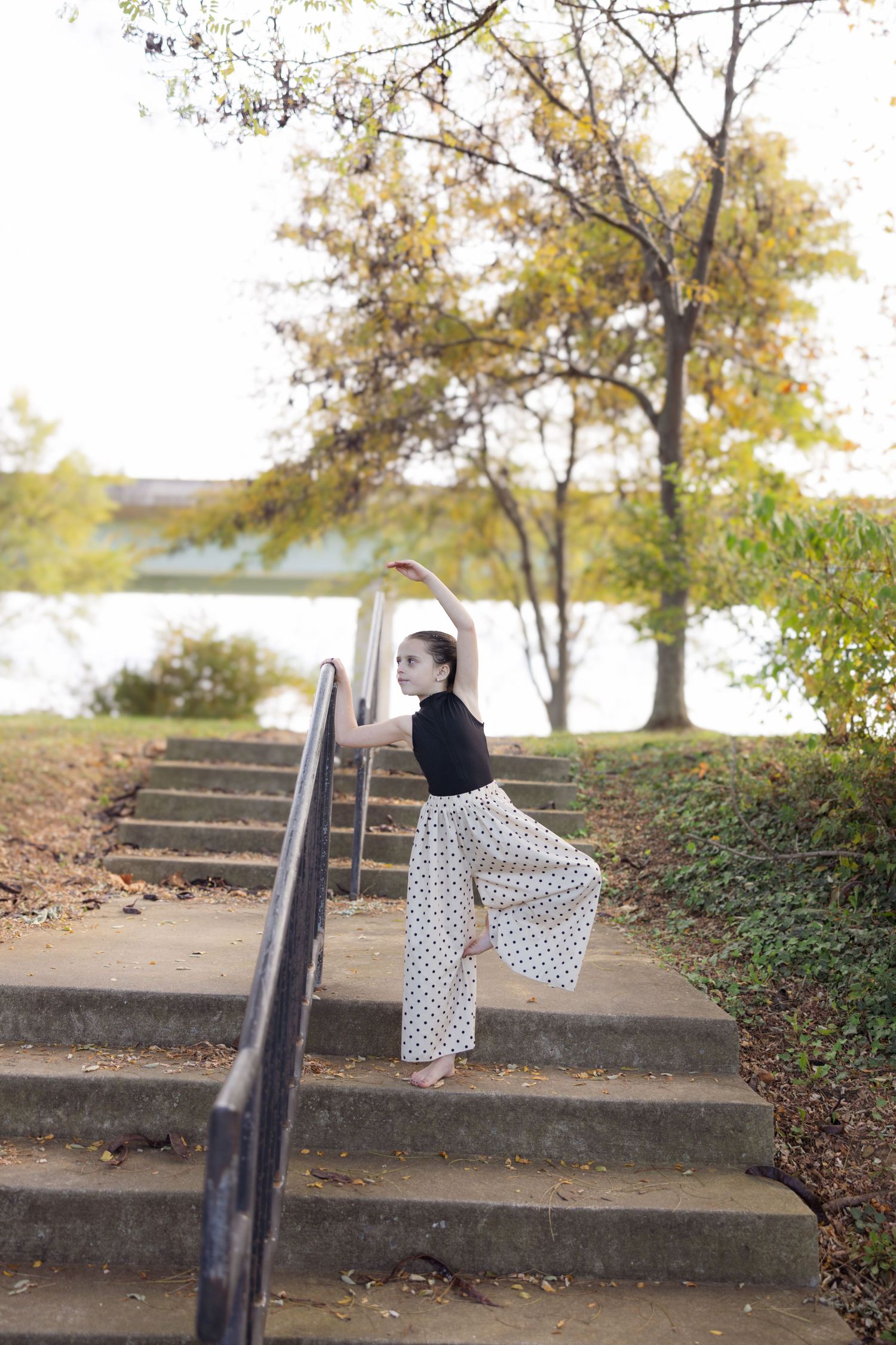 A person in a polka dot dress sits on concrete steps outdoors surrounded by autumn trees and foliage.