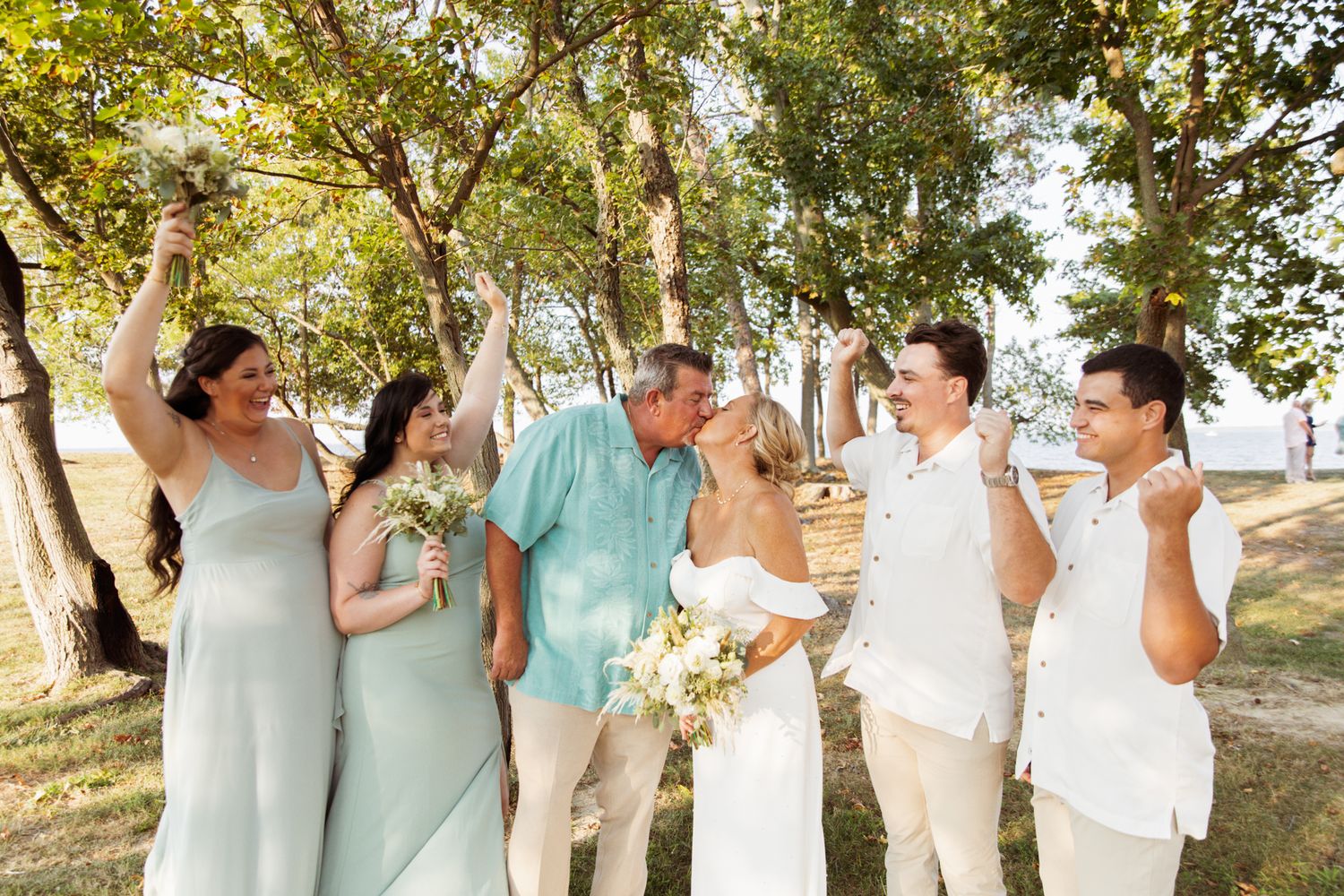 A joyful wedding party celebrates outdoors under trees, with bridesmaids in sage green and groomsmen in white.