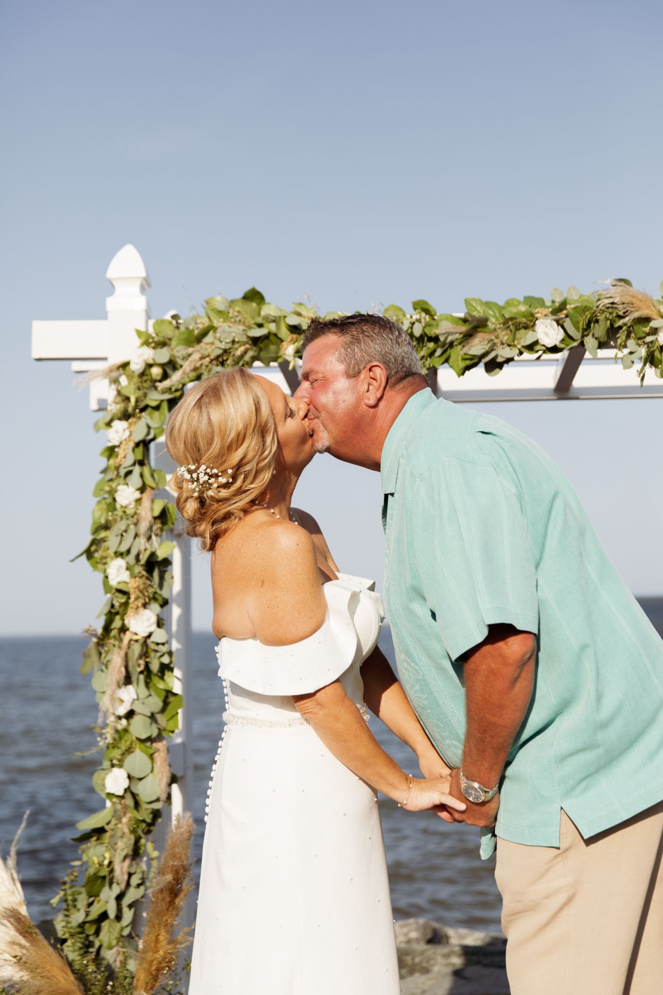 Couple sharing a kiss at outdoor beach wedding ceremony under white arbor decorated with lush greenery and white flowers.