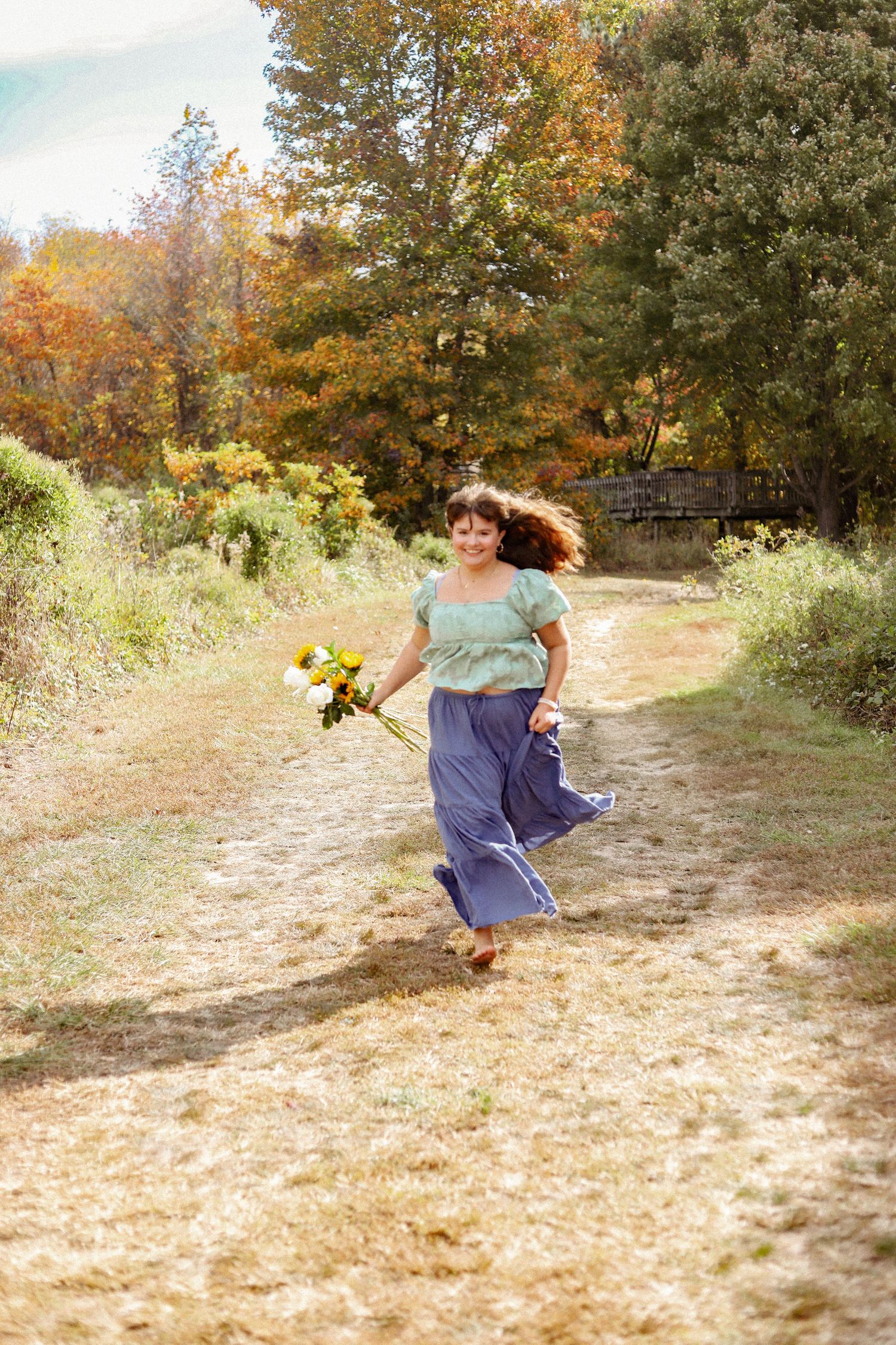 Person skips joyfully down a sunlit dirt path in autumn holding yellow flowers.