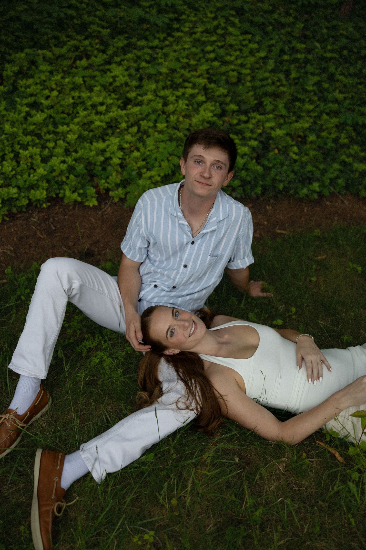 A couple relaxes on green grass, woman lying with head on man's lap, both wearing white outfits in a lush garden.