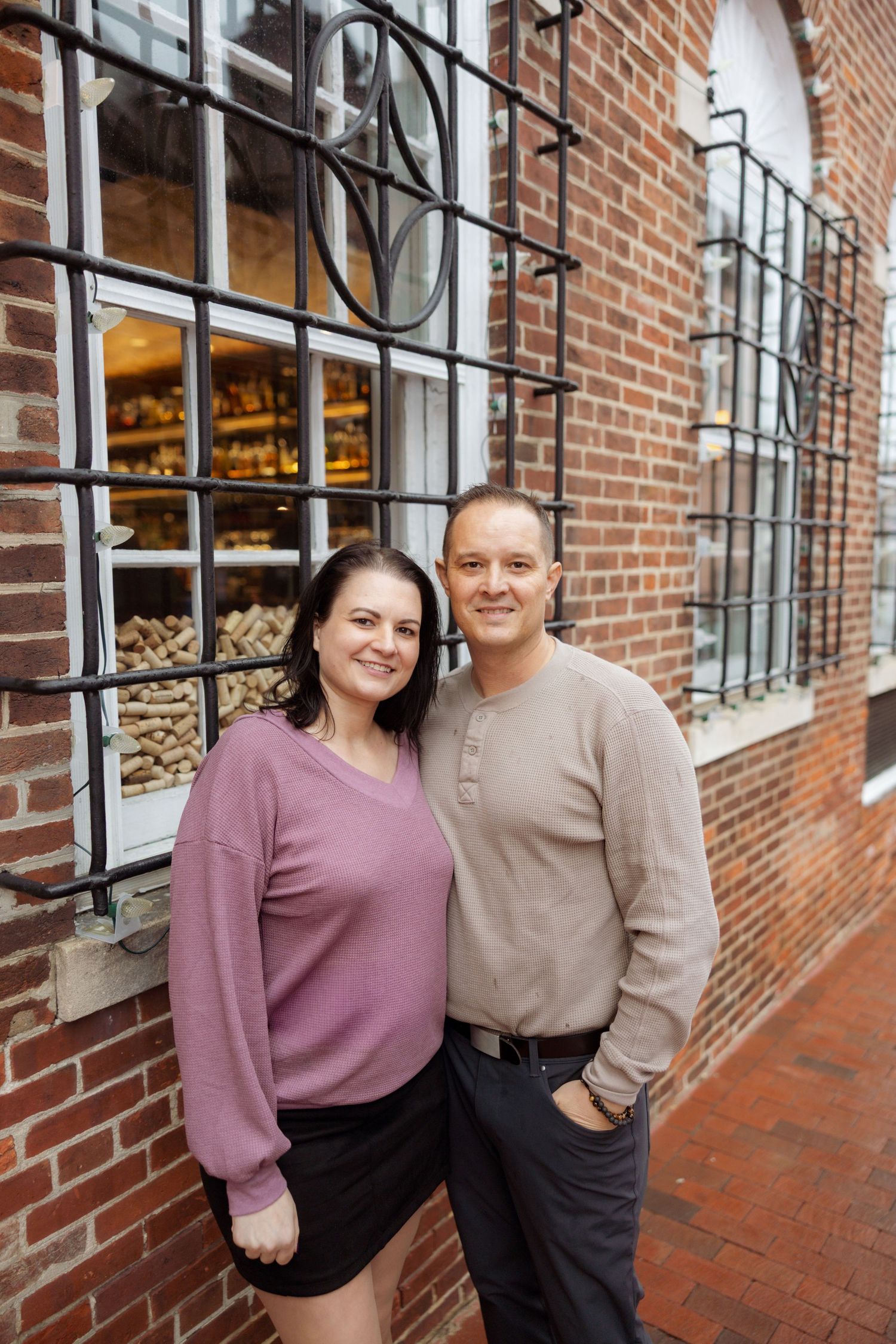 A couple stands smiling together outside a brick building with windows and decorative iron bars.