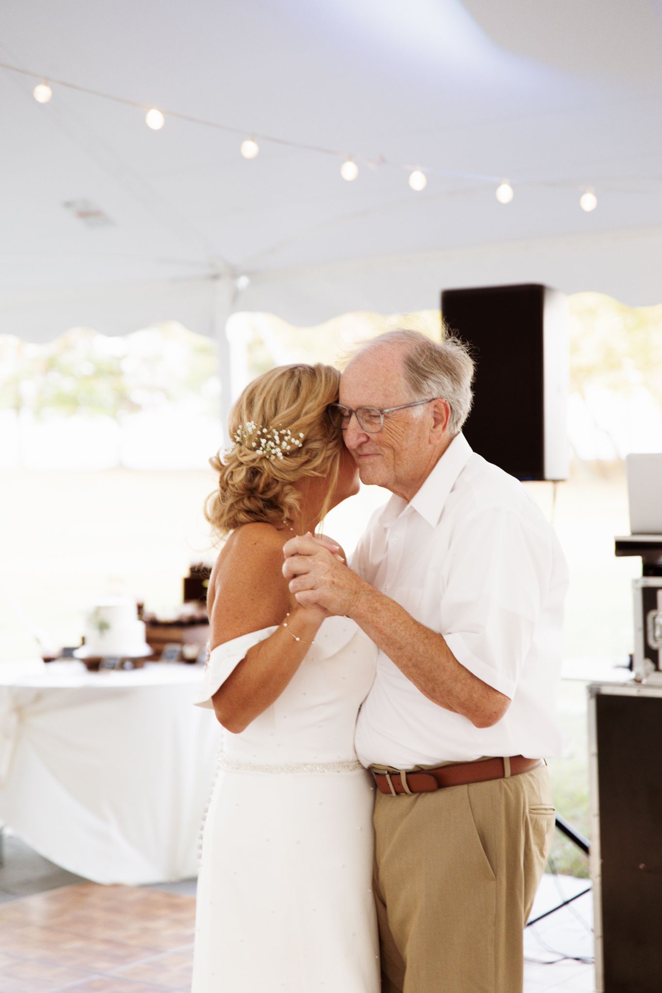 Elderly couple sharing a tender dance at a wedding reception under a white tent with string lights.