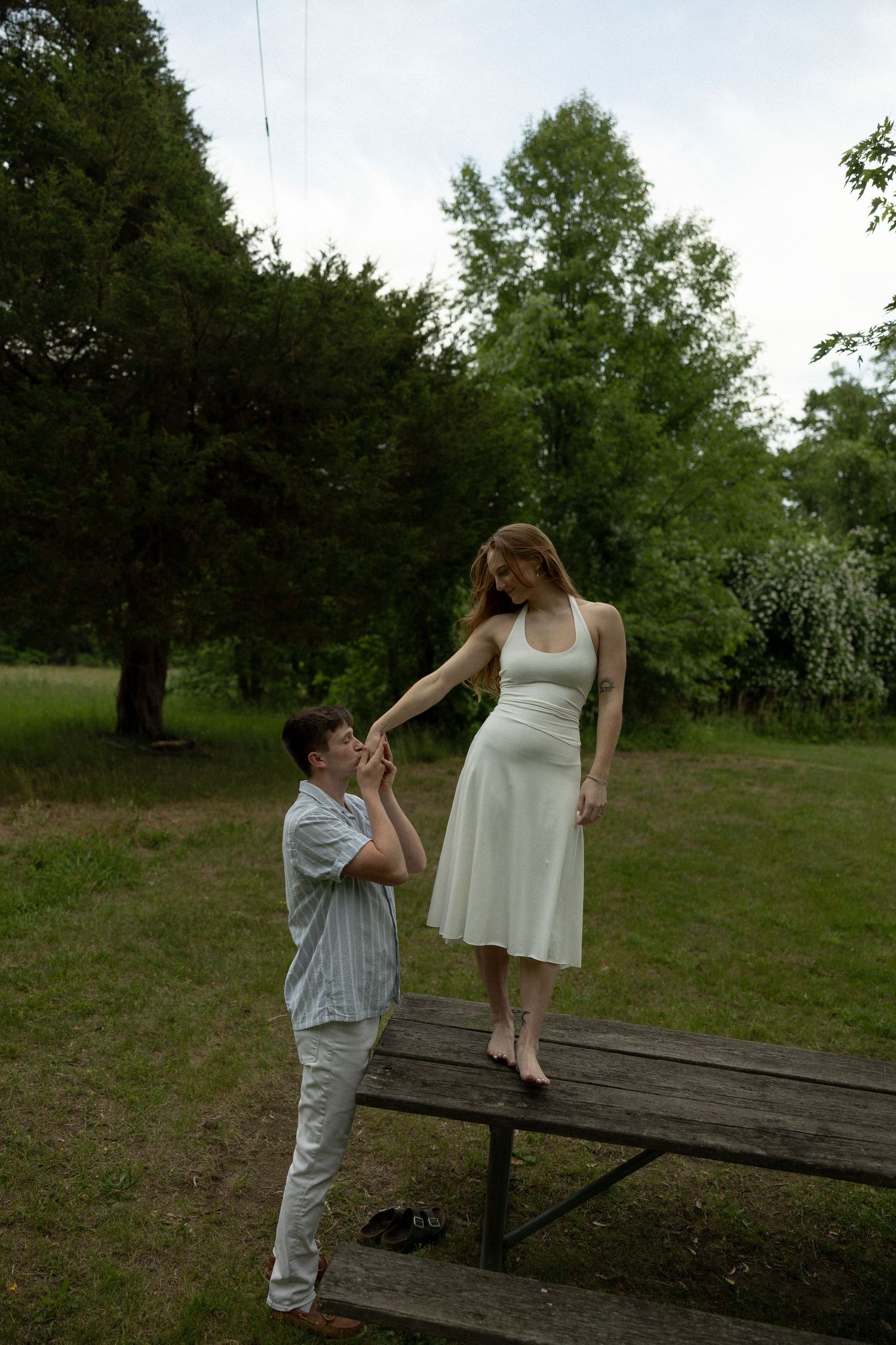 A man kisses a woman's hand as she stands on a picnic table wearing a white dress in a lush green park.