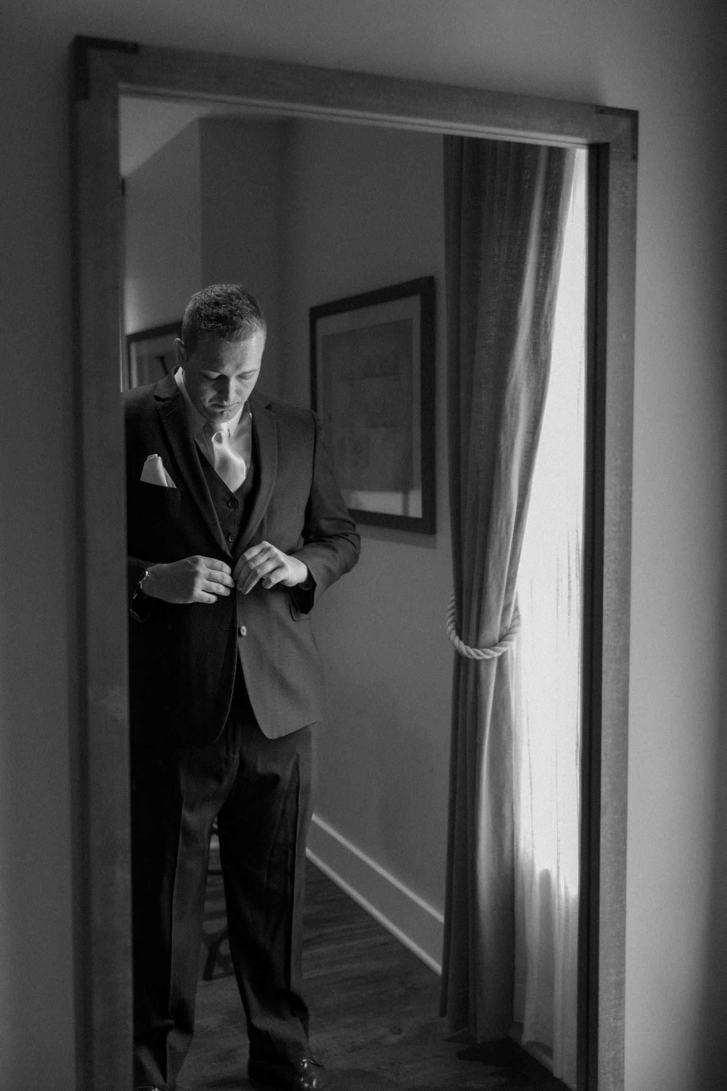 Groom buttoning his suit jacket in a mirror reflection, black and white wedding preparation photo.