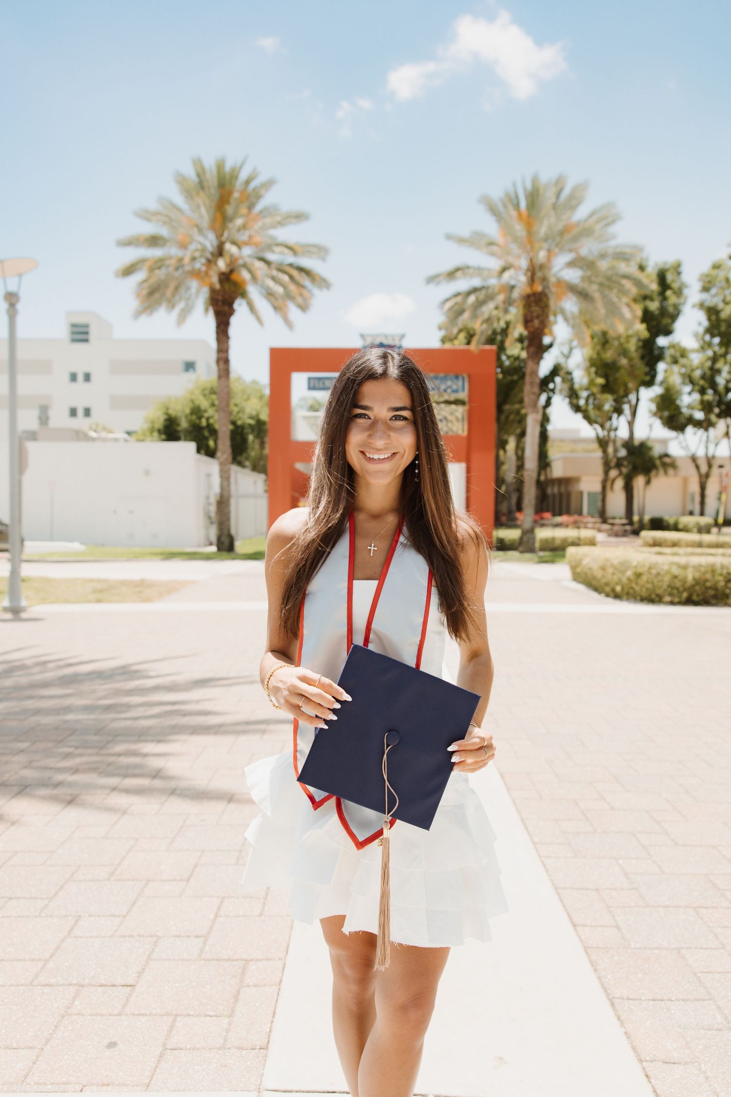 Graduate in white dress holds diploma on sunny campus walkway with palm trees.