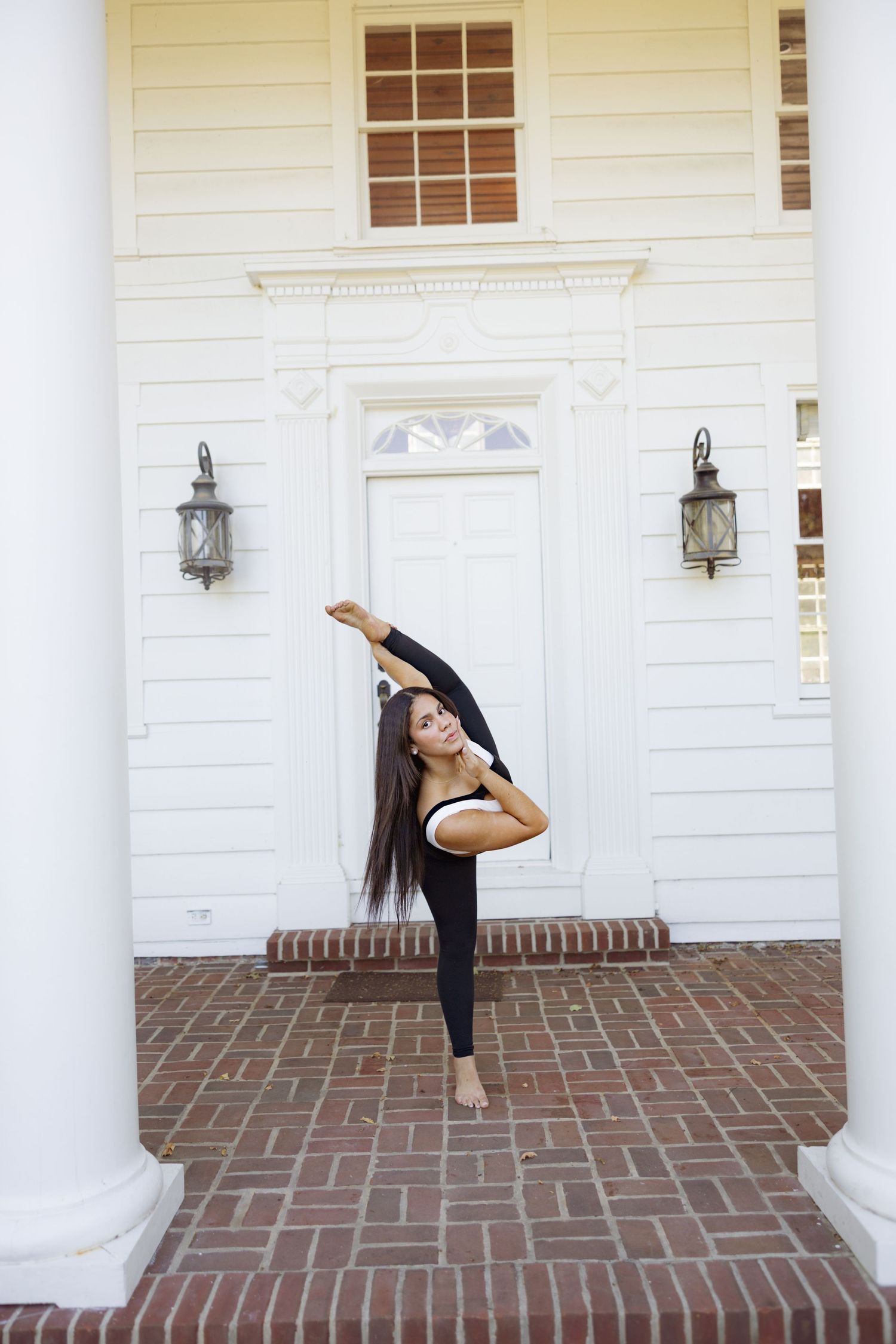 Dancer performs a high leg extension in front of a white colonial style building with brick porch and columns.