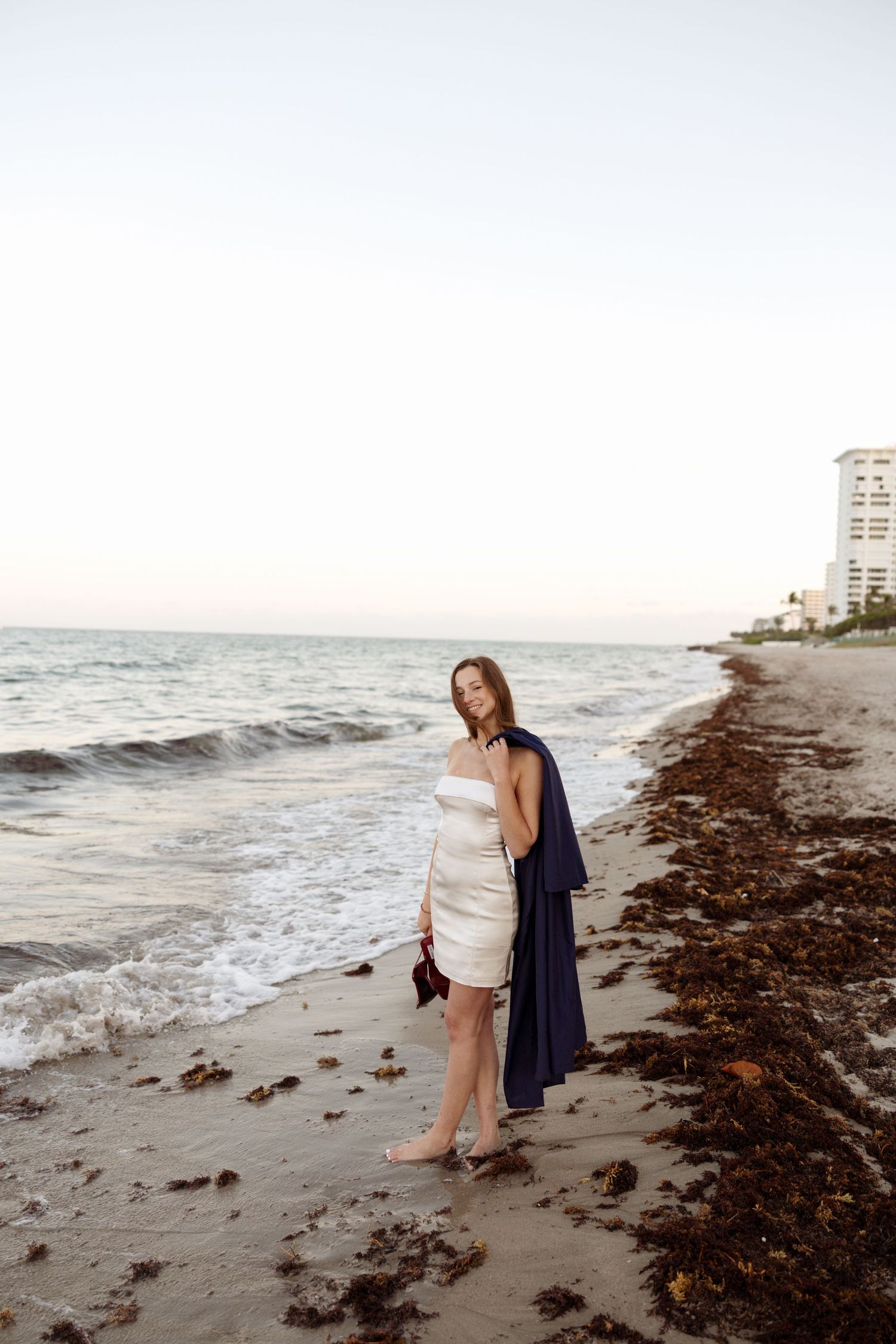 Person in white dress and navy cardigan stands on rocky beach at sunset.