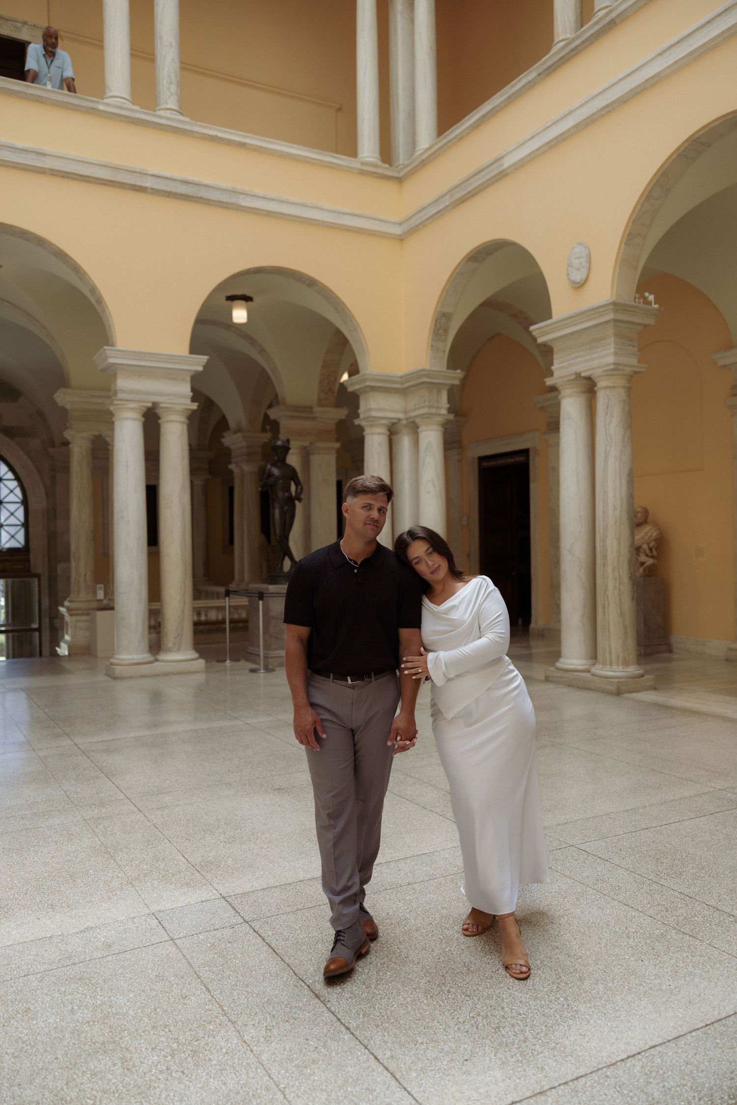 Two people walk through an elegant colonnade with cream-colored arches and marble floors in a historic building.