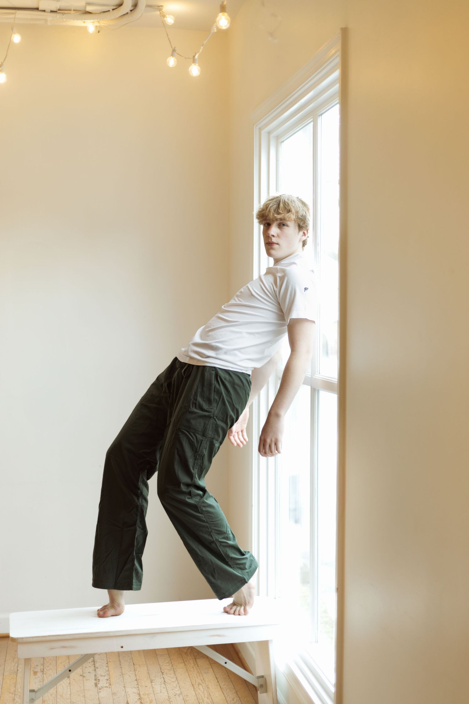 Person in casual attire poses artistically by bright window in minimalist setting.