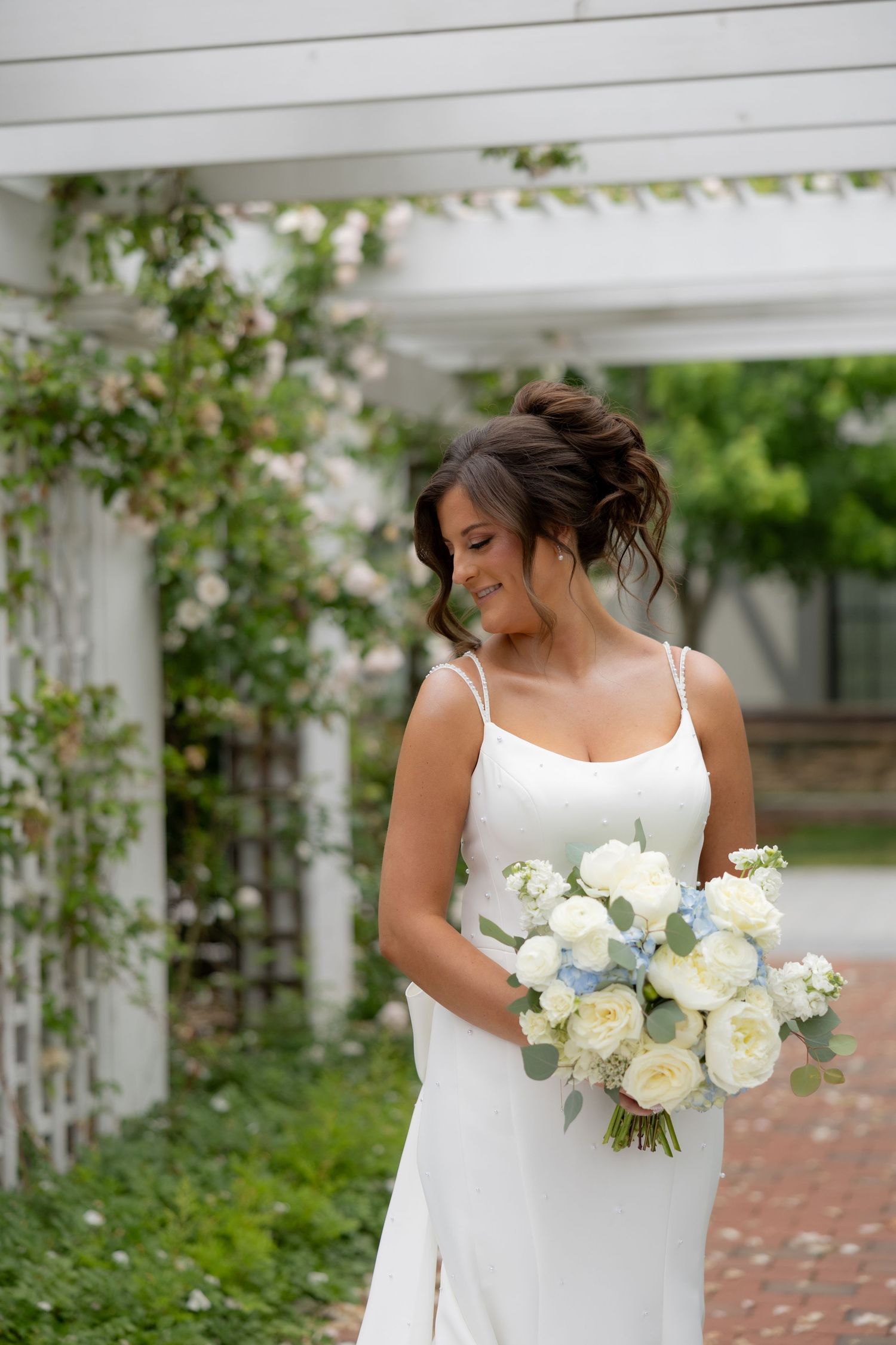 Bride in white spaghetti strap gown holding white and blue bouquet, smiling in a lush garden with white pergola.