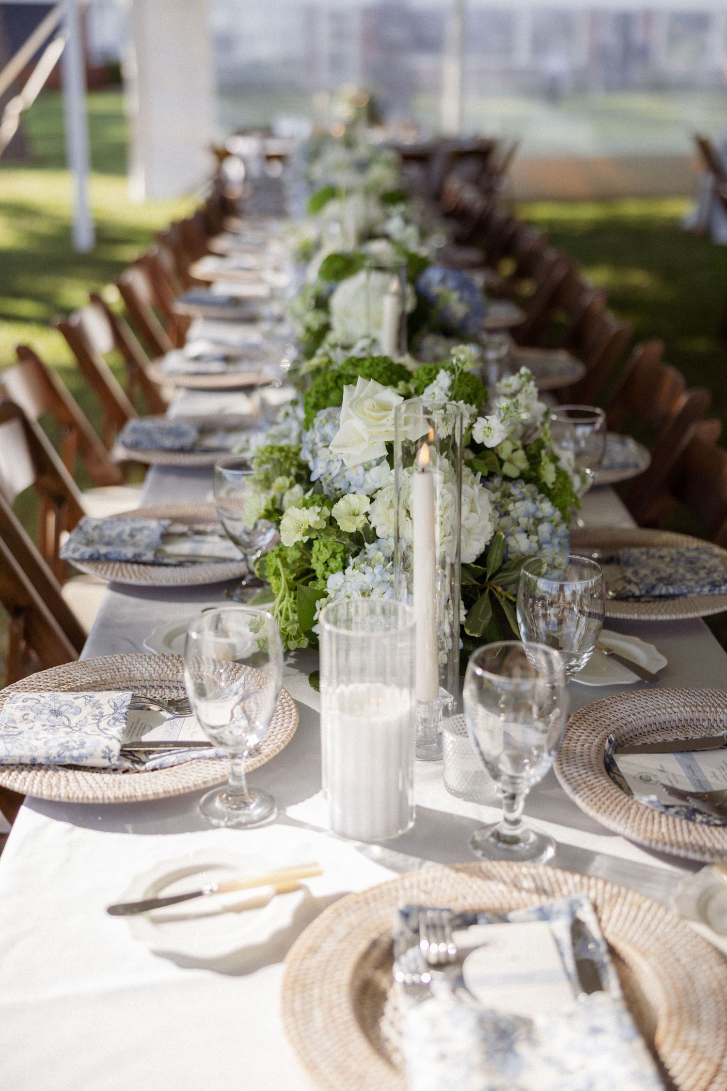 Elegant outdoor wedding reception table with white floral centerpieces, crystal glassware, and woven charger plates.