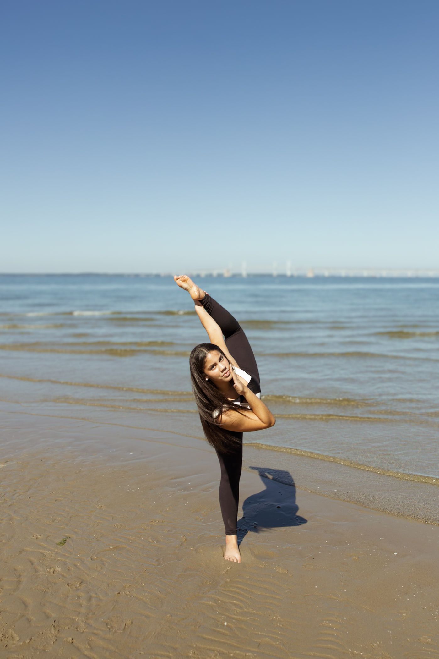 A dancer performs a graceful backbend pose on a sandy beach with calm ocean waters in the background.