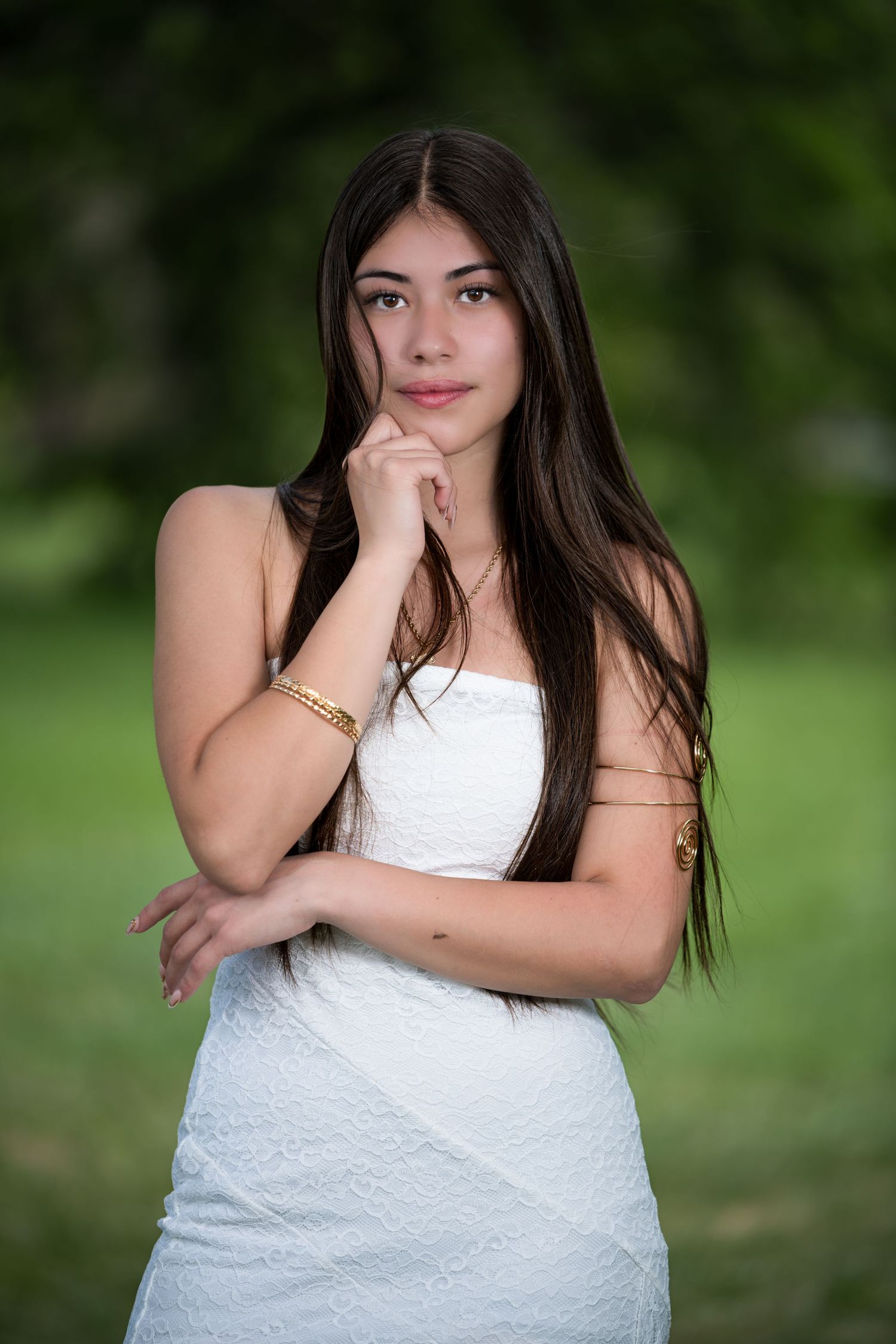 Young woman in white strapless dress with gold bracelets poses thoughtfully outdoors with green trees background.