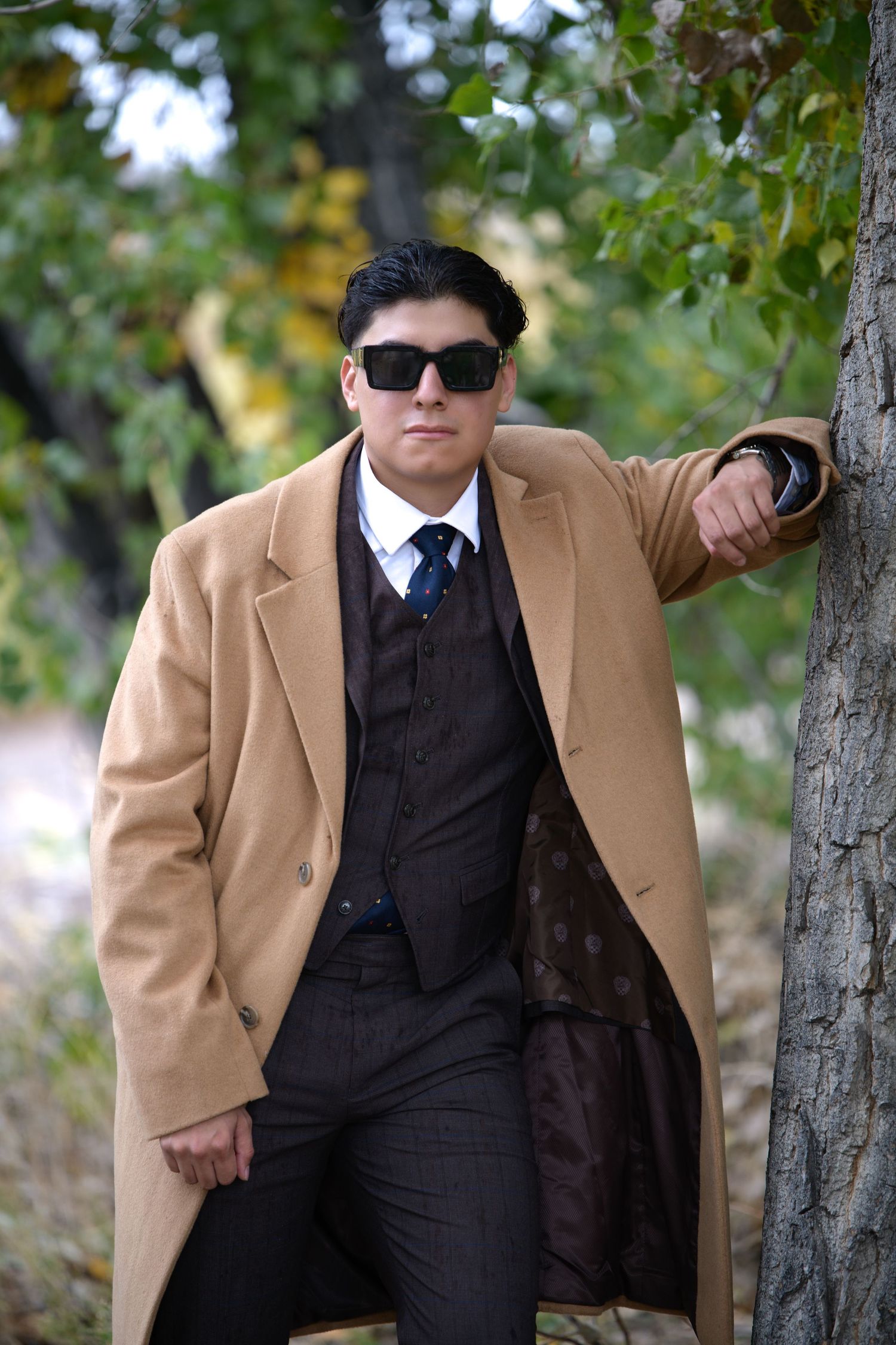 Man in camel overcoat, dark suit, tie, and sunglasses leaning against a tree in an autumn outdoor setting.