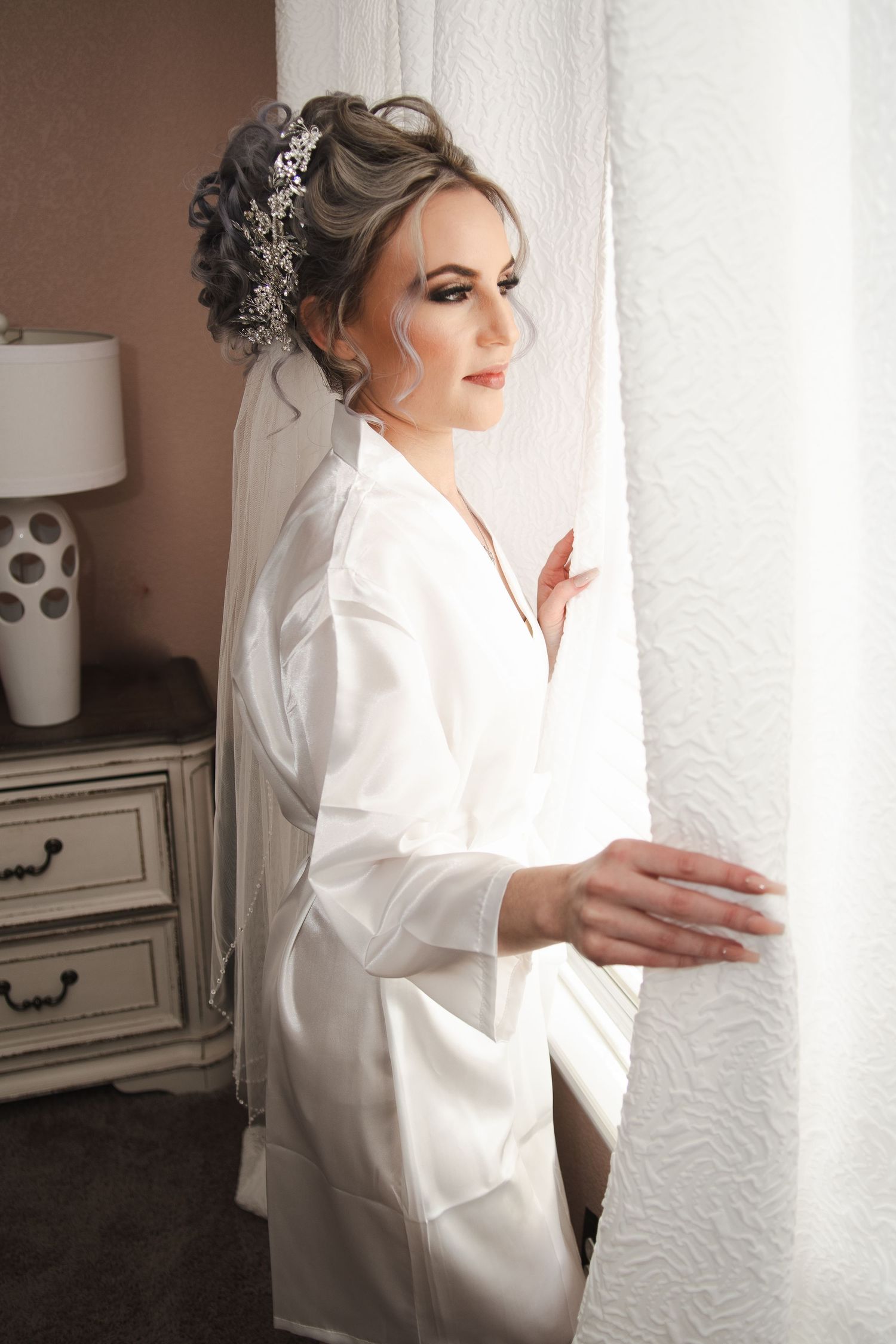 Elegant bride in white satin robe with updo hairstyle and veil looking out window before wedding.