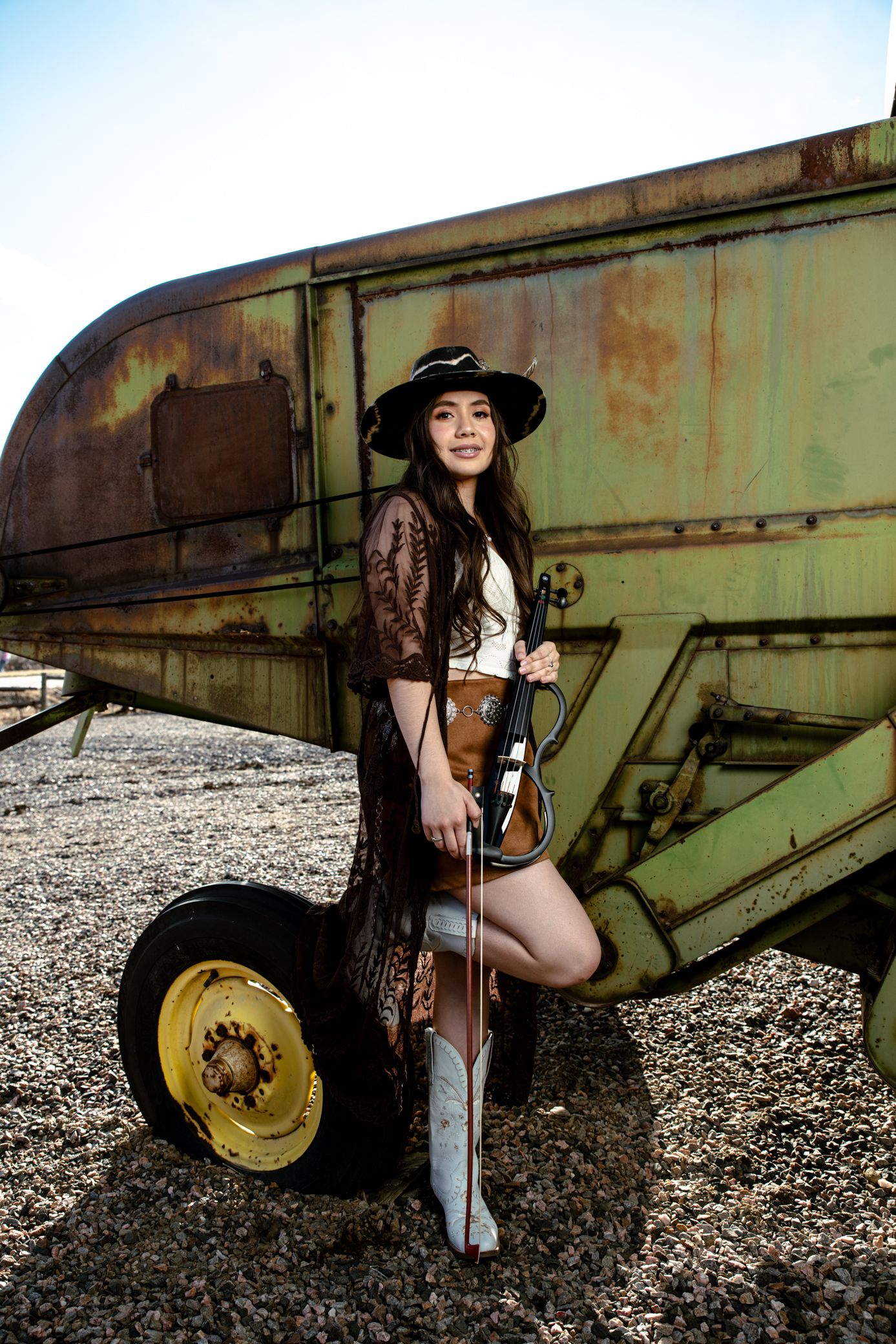Woman in cowboy hat holding violin posing against rustic green farm equipment outdoors.