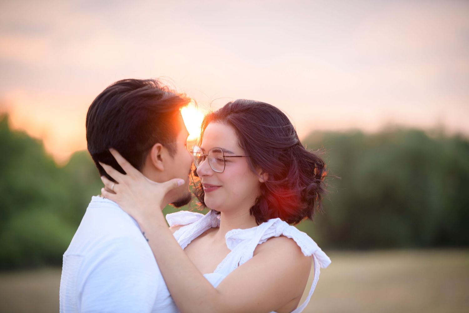 Couple sharing a romantic moment at sunset in a field, woman with red hair wearing glasses embracing her partner.