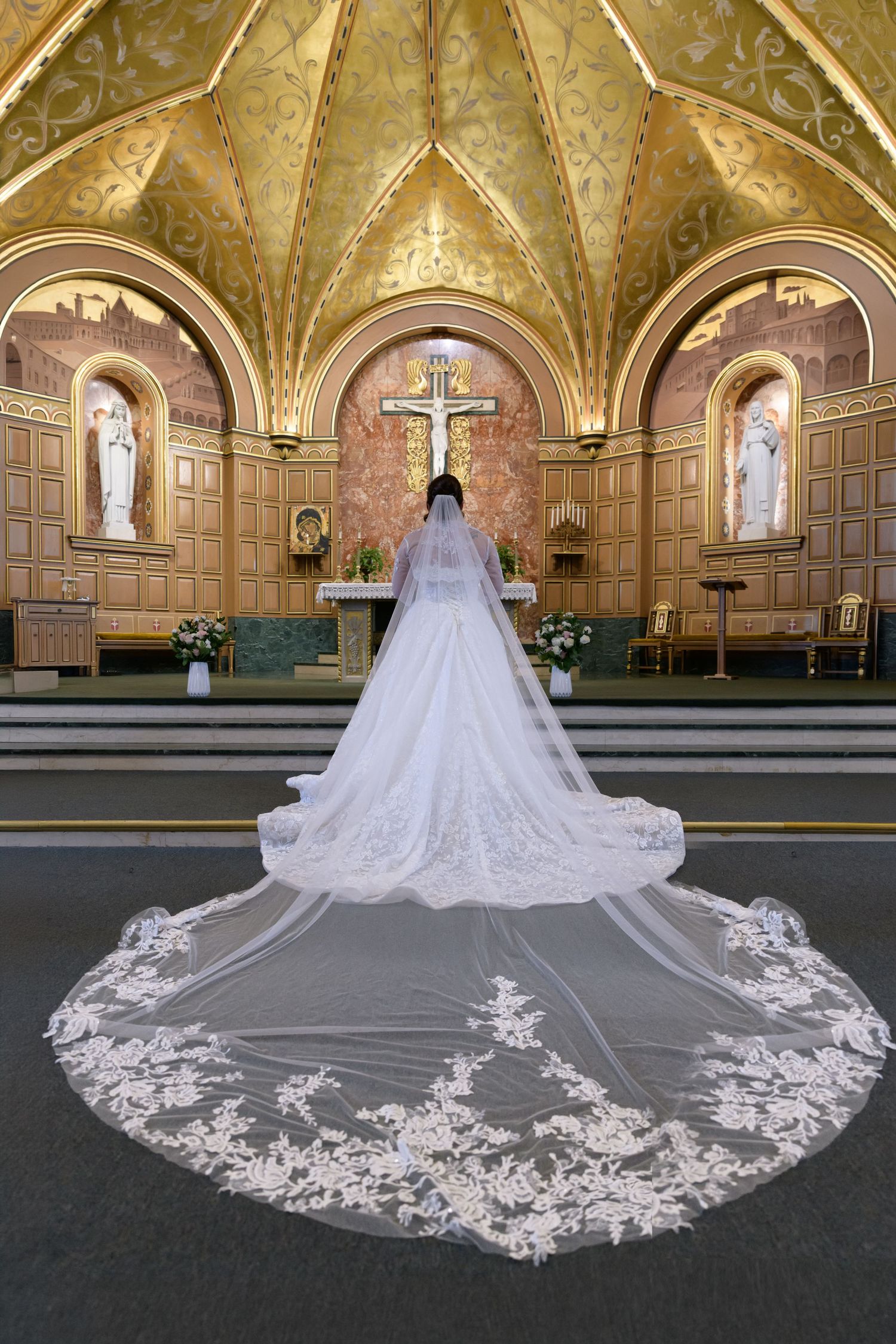 Bride in white lace wedding gown with cathedral veil stands before ornate golden altar in elegant church.