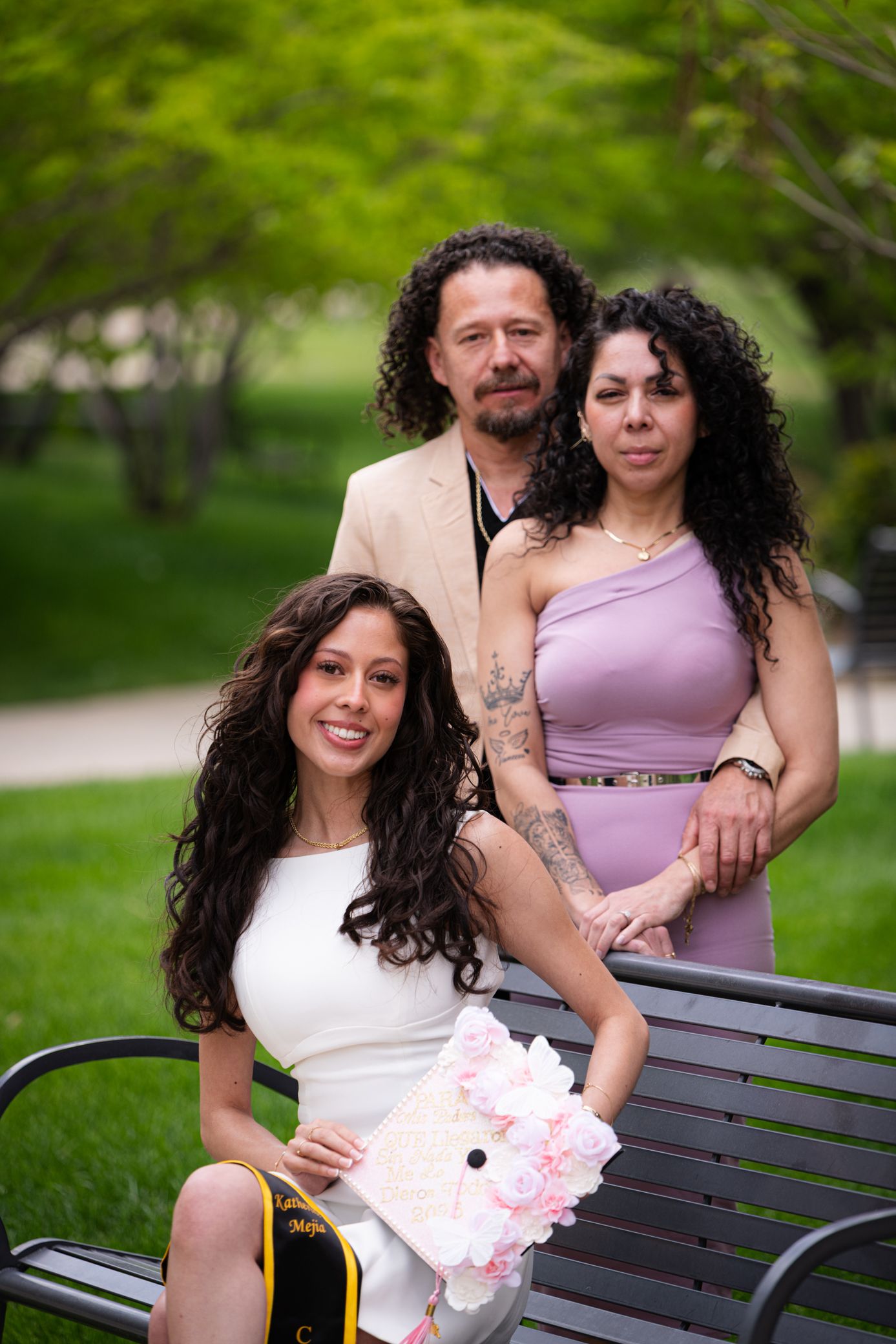 A family of three poses in a park, with a smiling graduate in white holding a decorated cap and pink flowers.