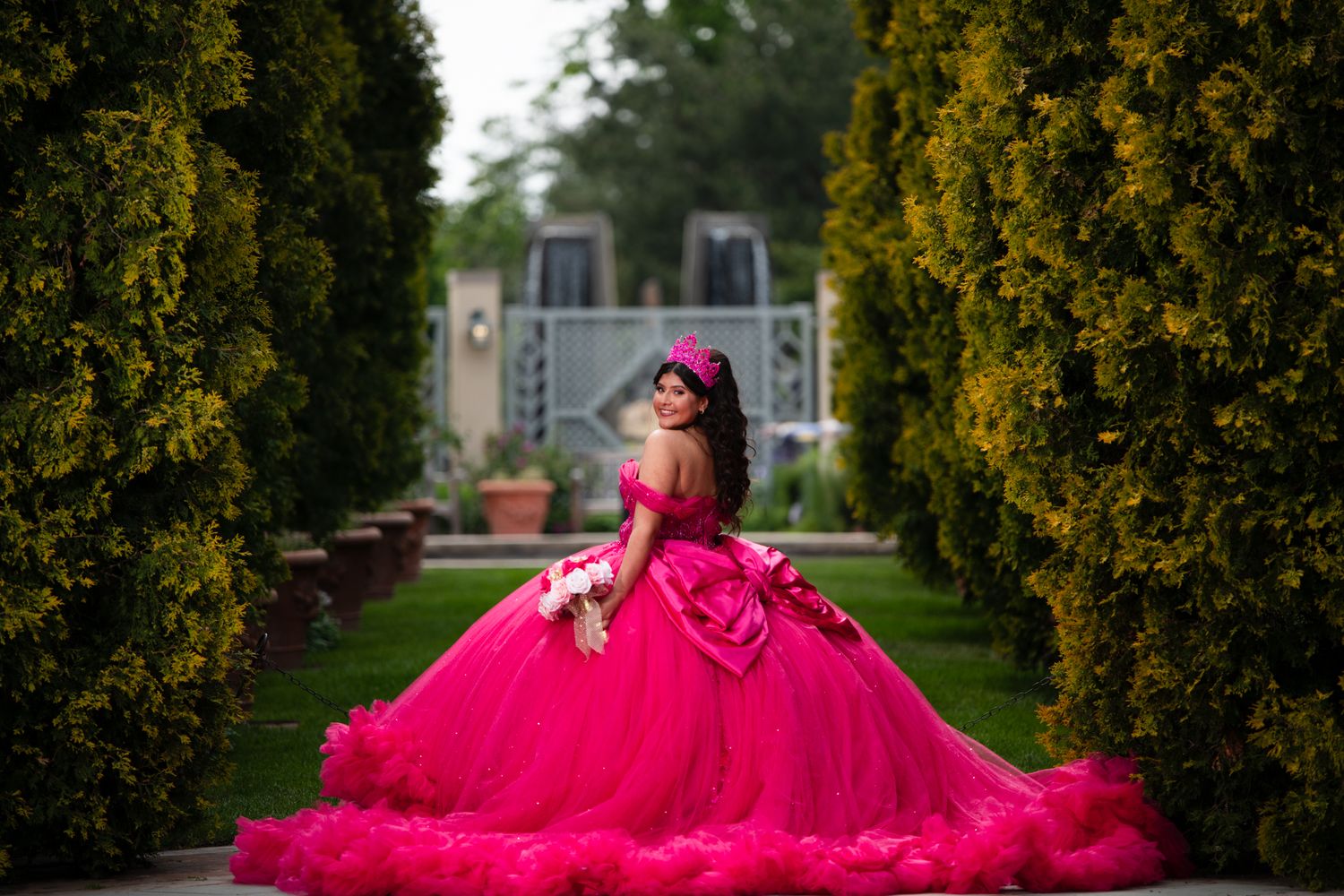 Young woman in vibrant hot pink quinceañera ball gown poses between tall hedges in a formal garden.