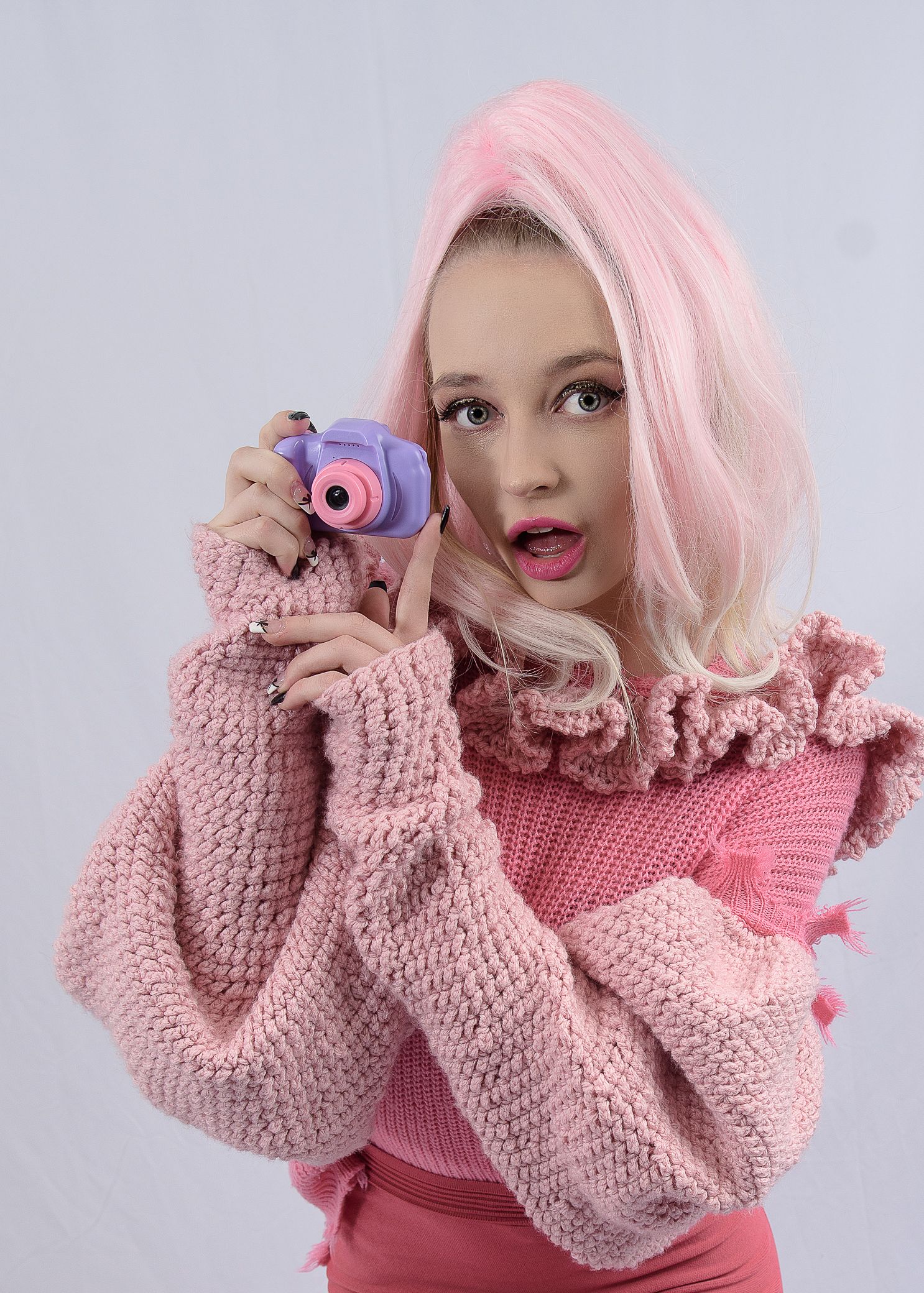 Young woman with pink hair holding a purple toy camera wearing a pink knit sweater against white background.