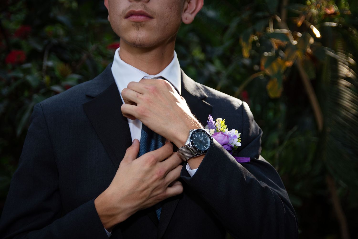 Groom adjusting his tie wearing a dark suit with floral boutonniere and luxury watch at wedding.