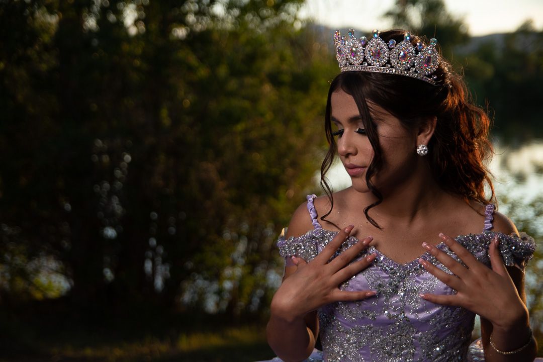 A young woman in a sparkling lavender quinceañera gown and tiara poses outdoors at golden hour.