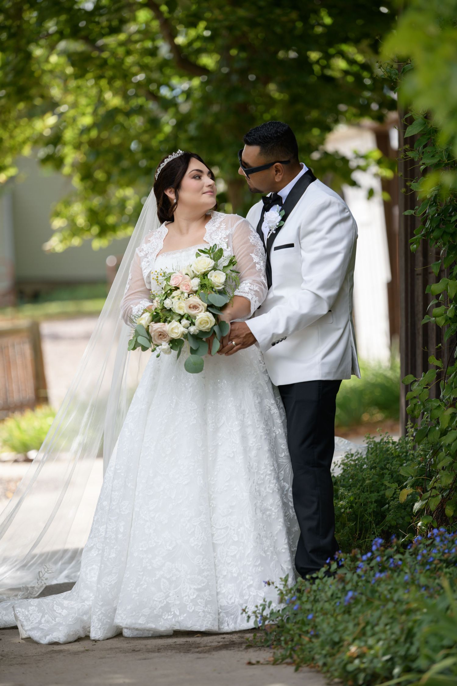 Bride in lace gown holding bouquet stands with groom in white tuxedo jacket outdoors among green trees.
