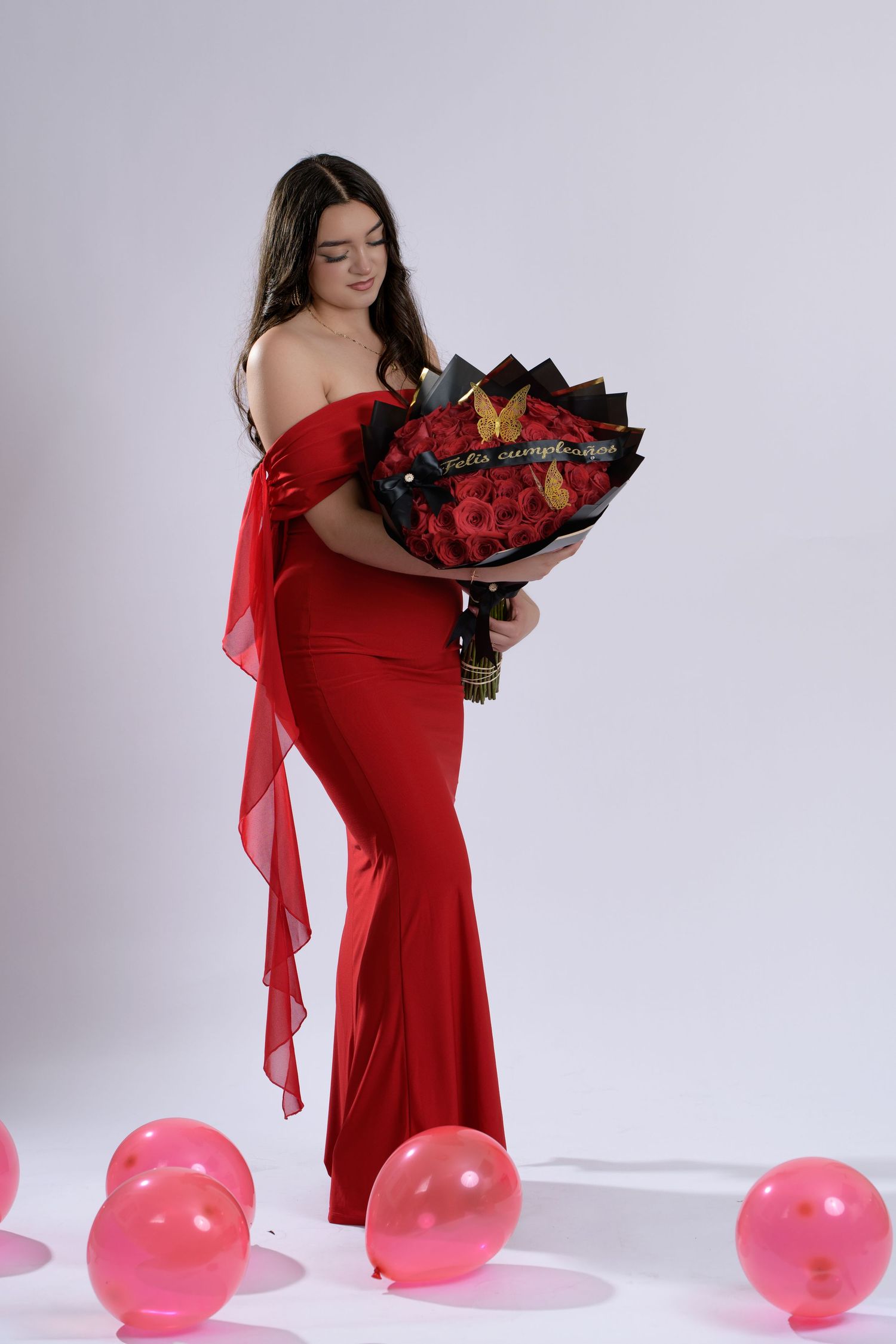Young woman in red off-shoulder gown holding large red rose bouquet, surrounded by pink balloons on white background.