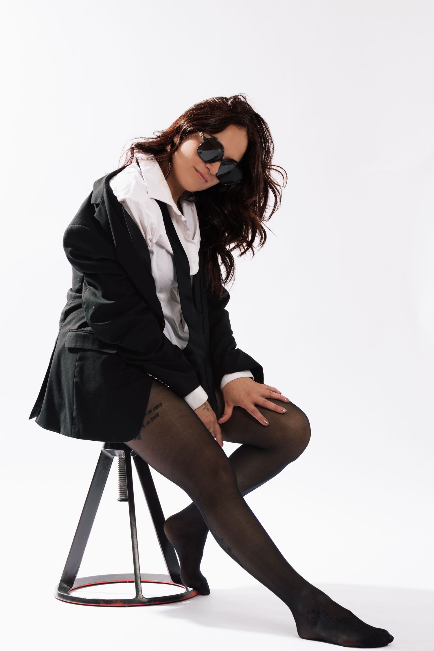Woman in black blazer and stockings sits on stool wearing heart-shaped sunglasses in studio.