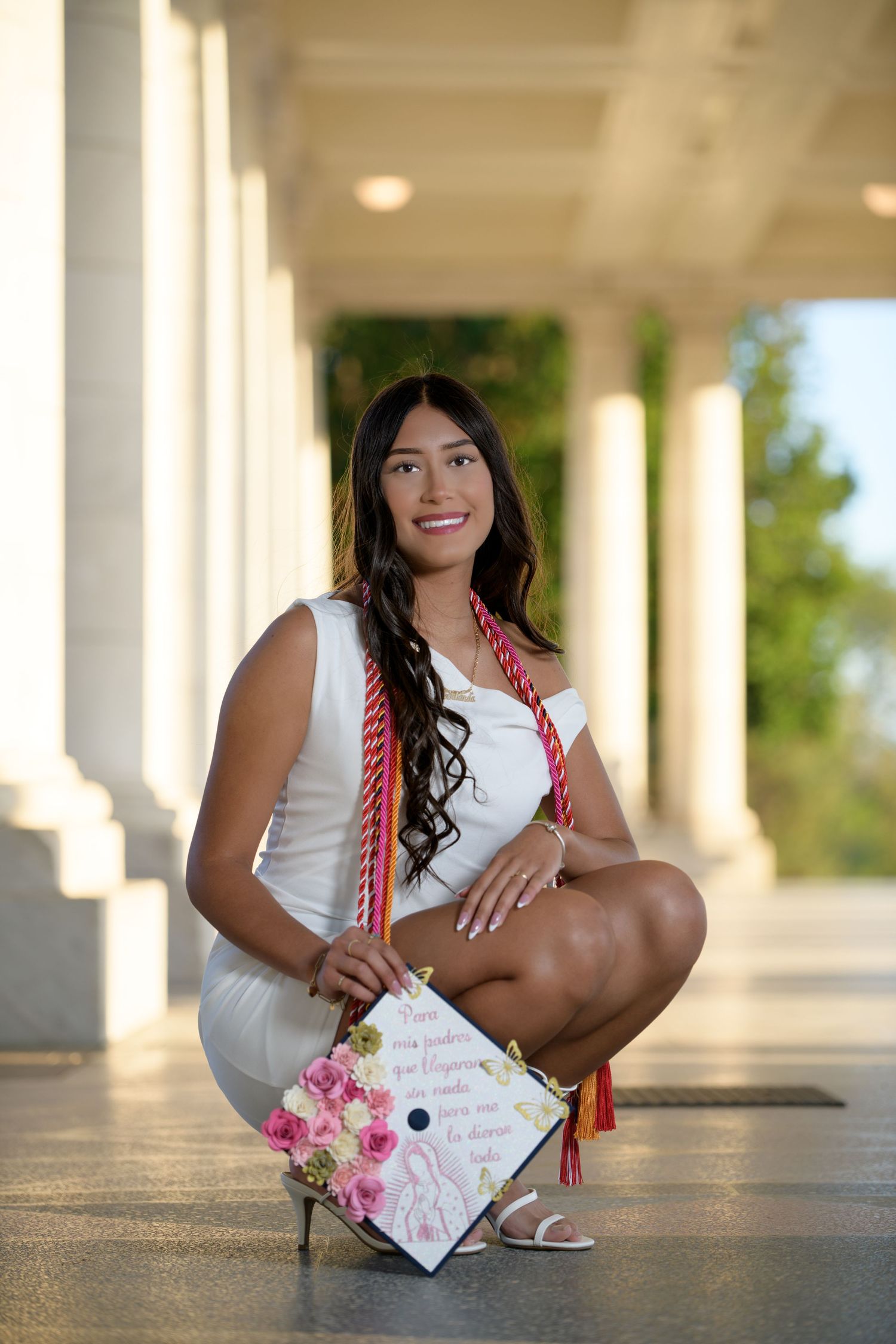 Smiling graduate in white dress holds decorated cap with flowers at columned outdoor venue.