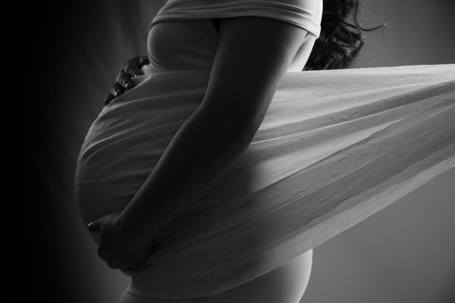 Artistic black and white silhouette of pregnant woman holding flowing white fabric over baby bump.