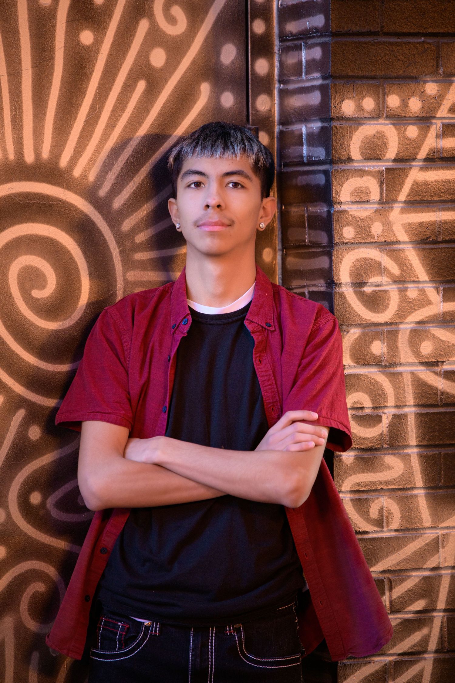 Young man in red shirt and black tee stands arms crossed against a brick wall with golden graffiti art.